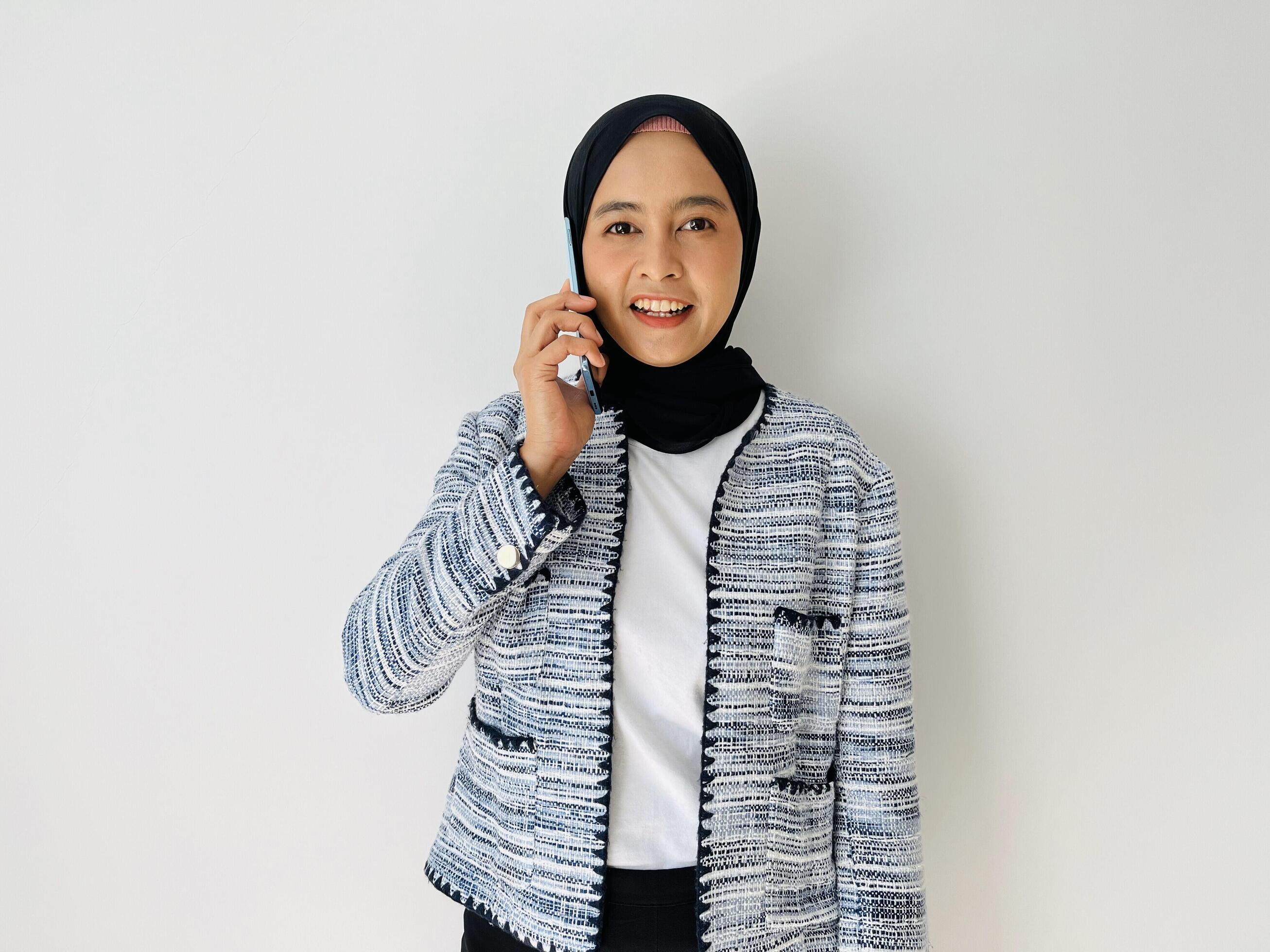 Young Asian woman smiling while using mobile phone having call. Standing female wearing black hijab and tweed blazer isolated on white background. Stock Free