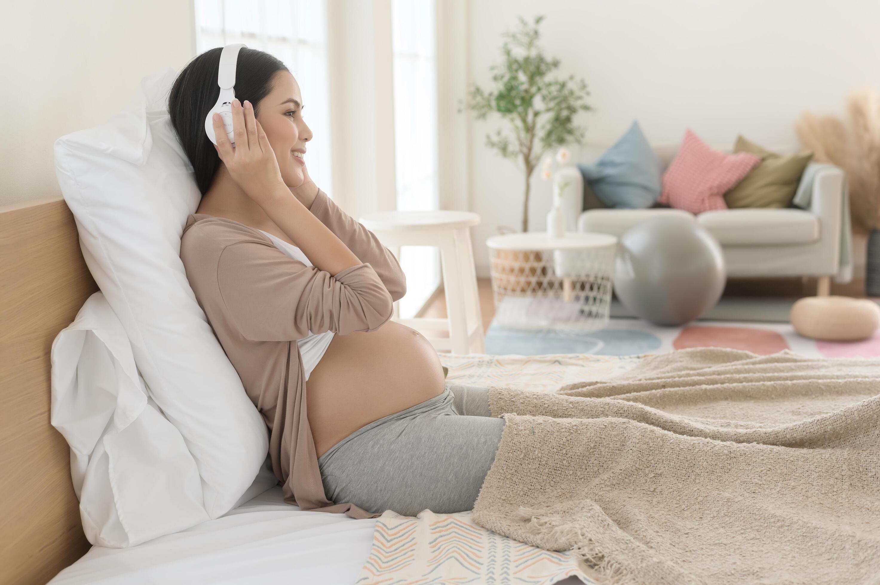 Happy pregnant woman with headphones listening to mozart music and lying on bed, pregnancy concept Stock Free