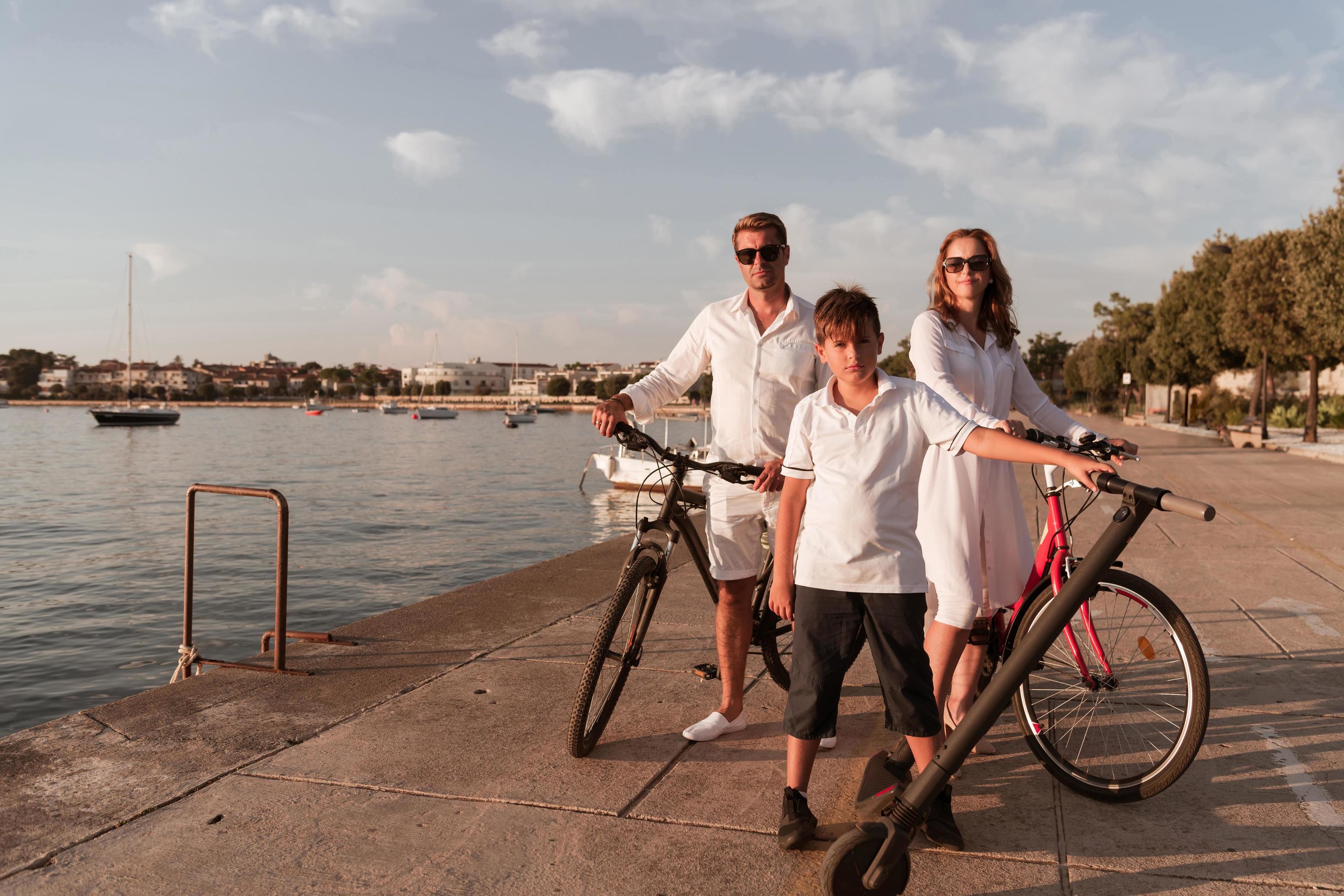 Happy family enjoying a beautiful morning by the sea together, parents riding a bike and their son riding an electric scooter. Selective focus Stock Free