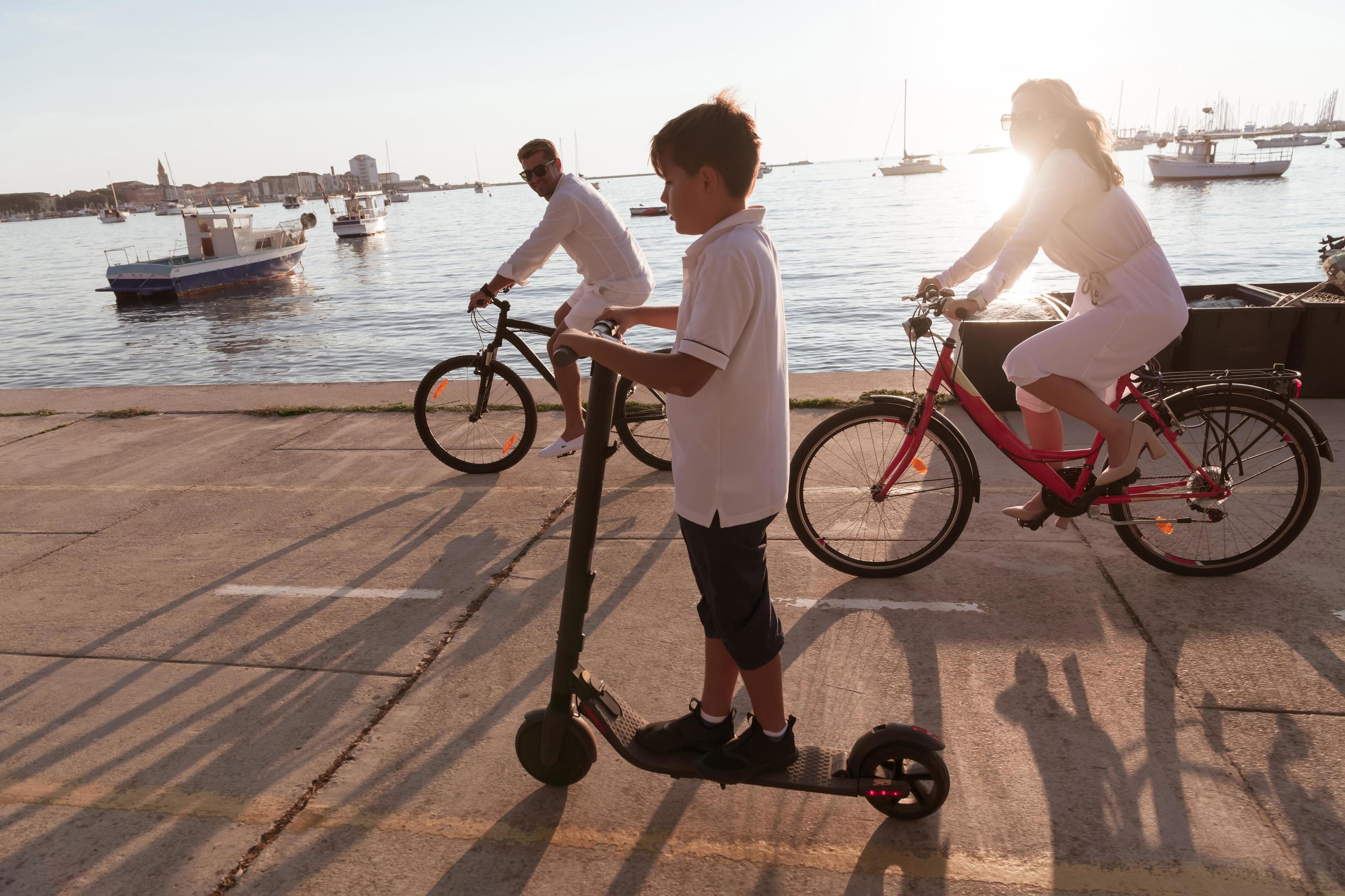 Happy family enjoying a beautiful morning by the sea together, parents riding a bike and their son riding an electric scooter. Selective focus Stock Free