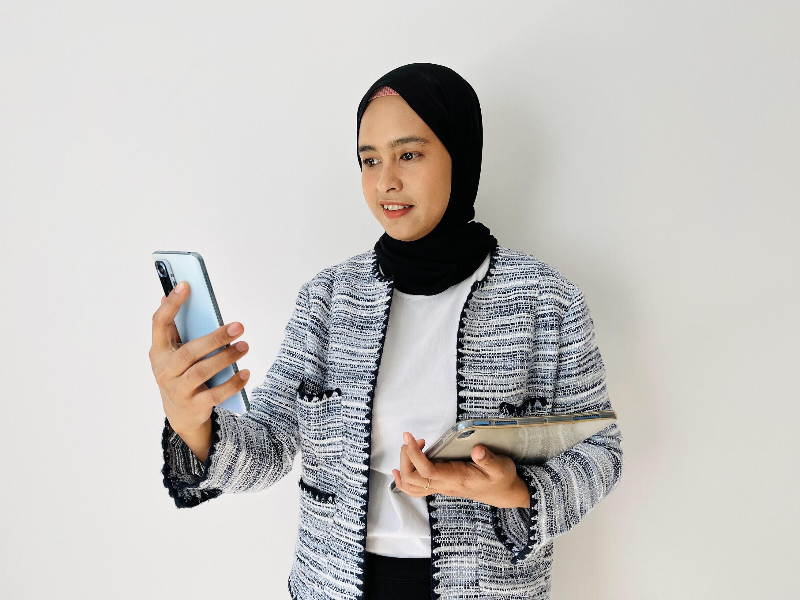 A young Asian woman is making a call for an online meeting using a mobile phone and holding a tablet Stock Free