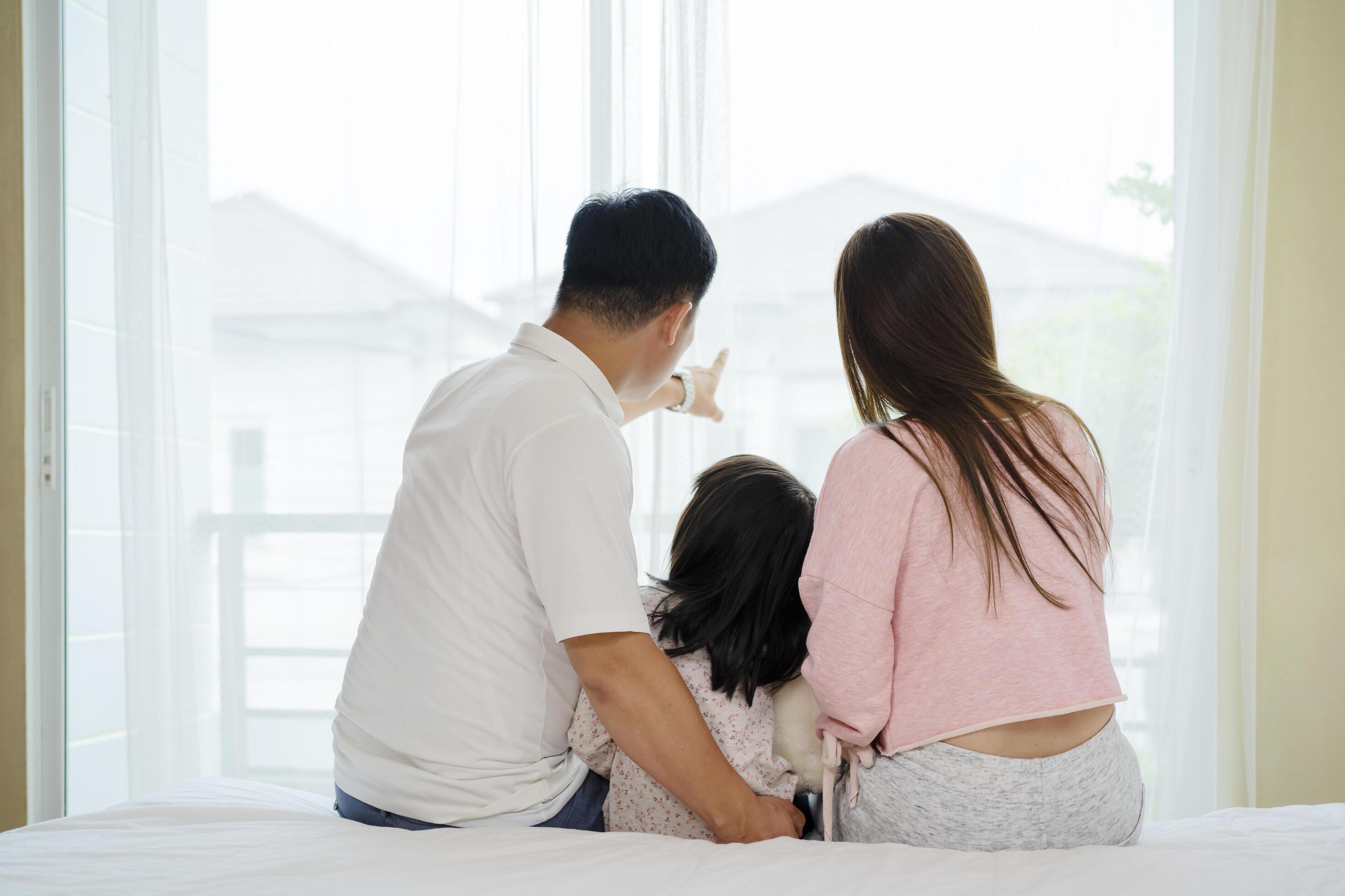 A happy Asian family, parents, daughter together Sitting on the bed looking out the window Stock Free