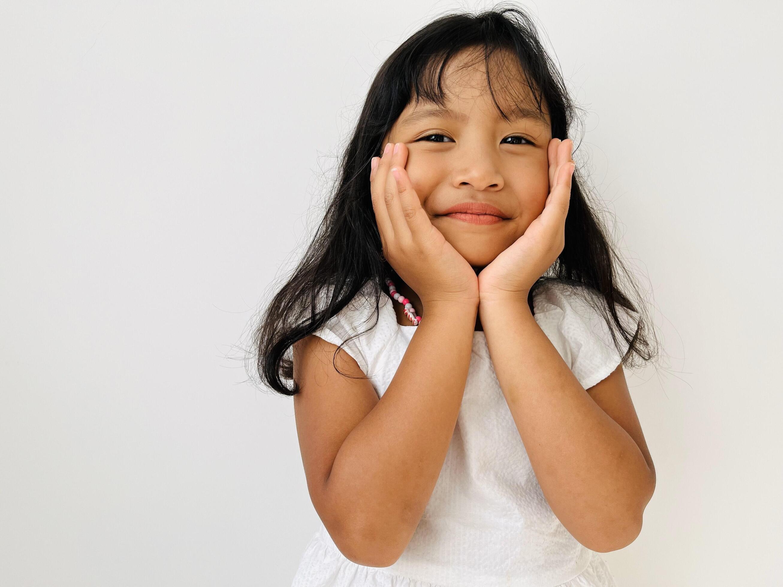 A cute 5 year old little girl wearing a white dress smiles with both hands holding her chin Stock Free