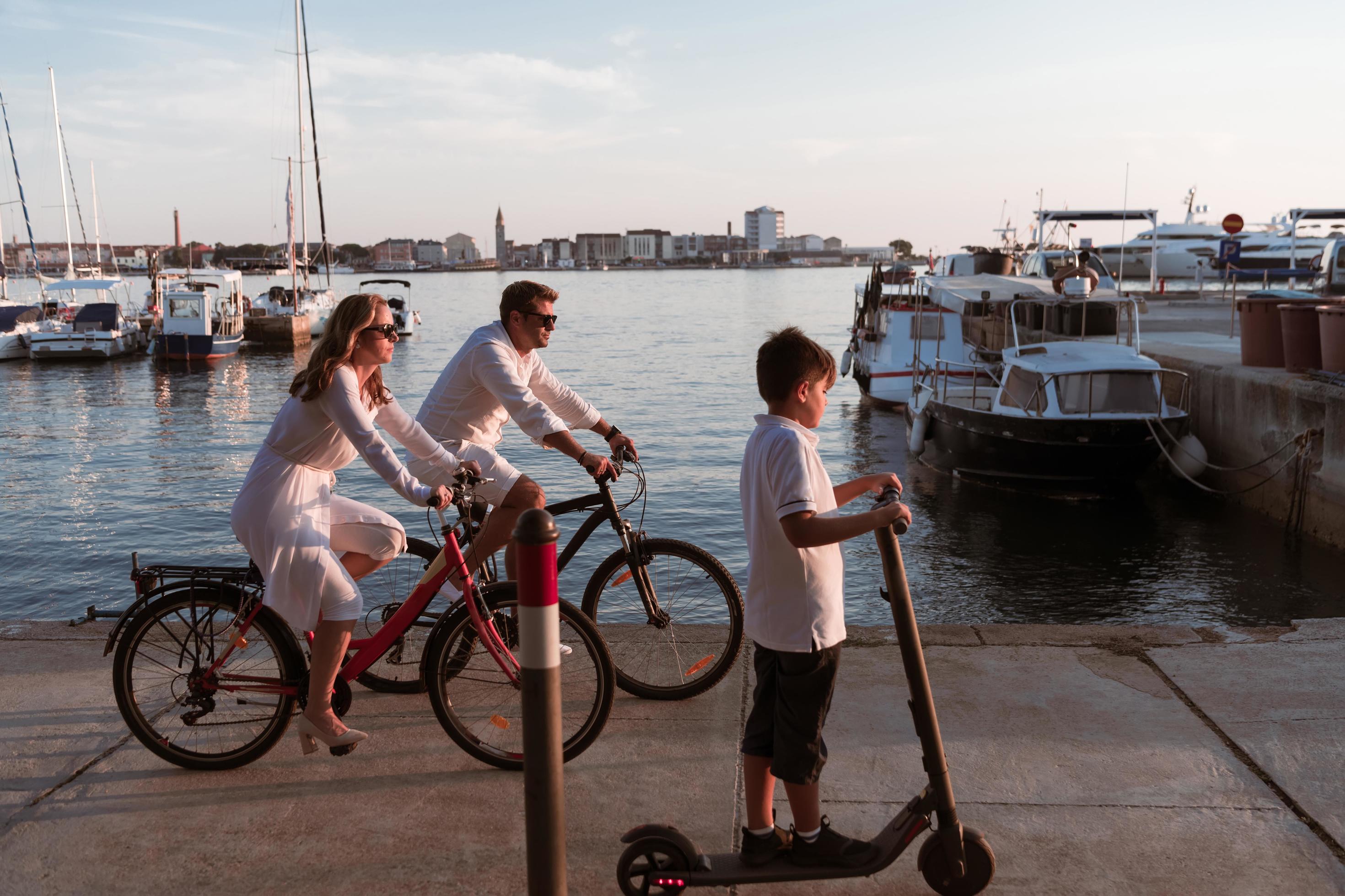 Happy family enjoying a beautiful morning by the sea together, parents riding a bike and their son riding an electric scooter. Selective focus Stock Free