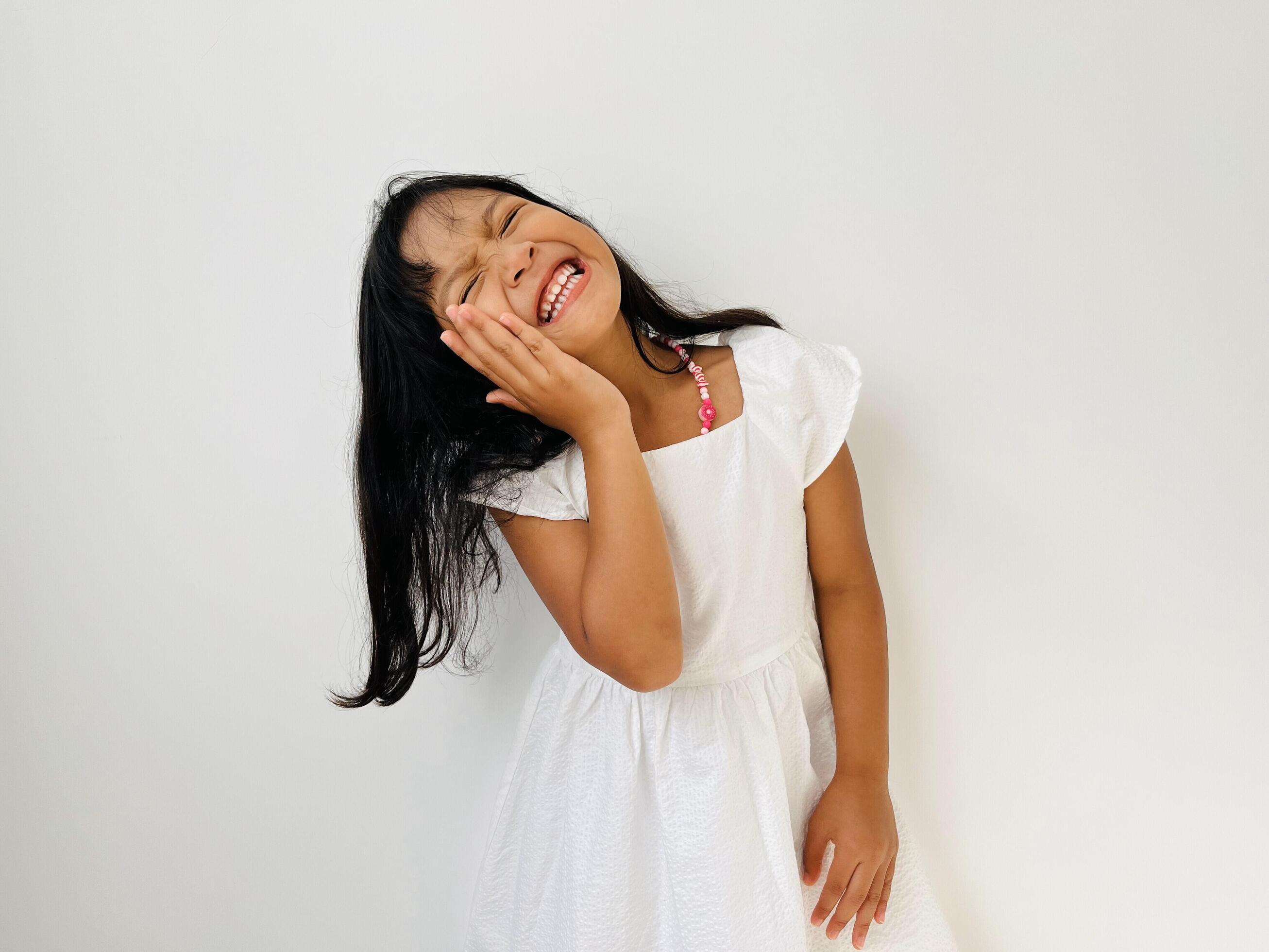 A five-year-old Asian girl smiles broadly with her eyes closed and one hand on her cheek Stock Free