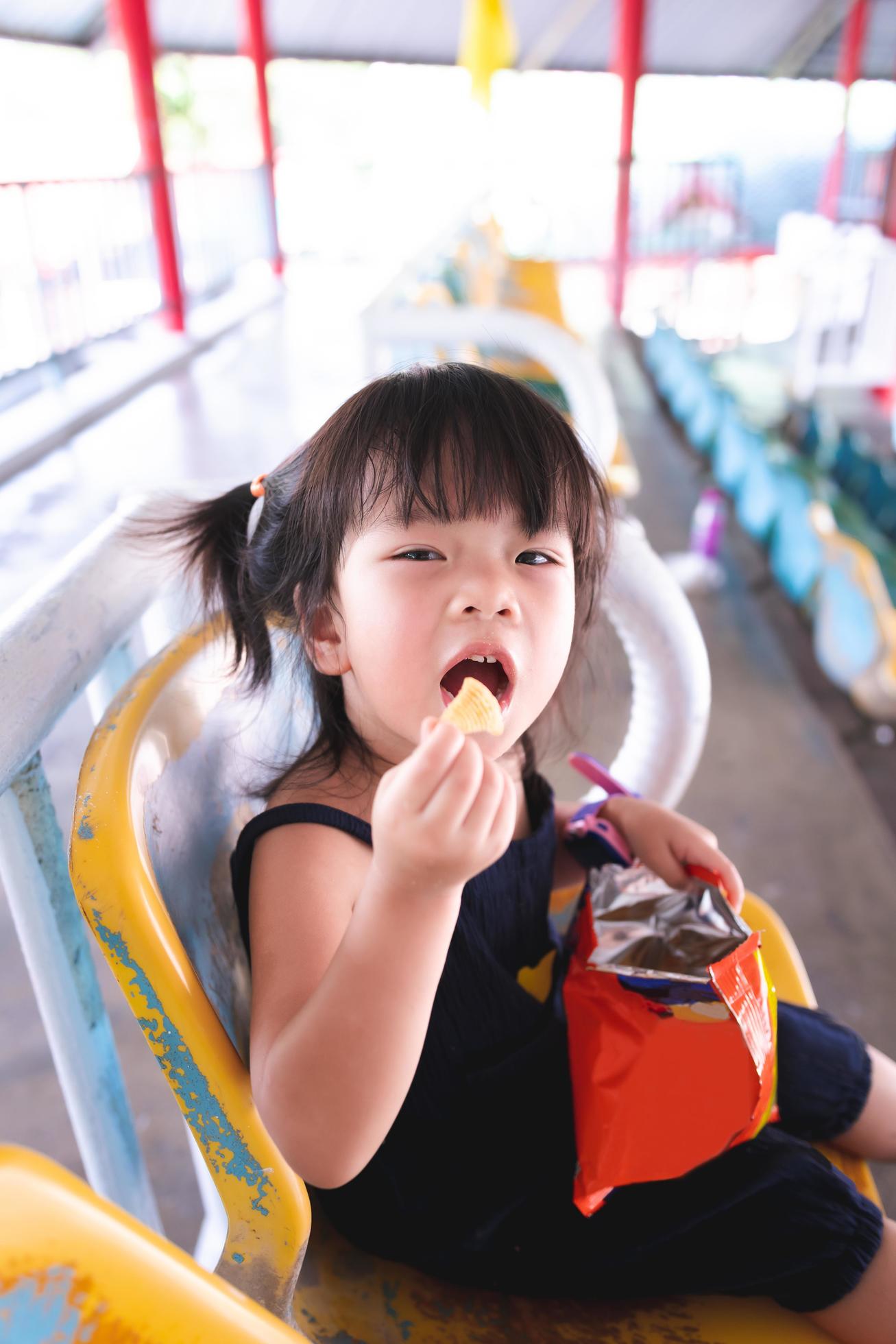 Happy cute 3 year old kid girl eating snacks on grandstand. Child picks up crunchy food in her mouth. Vertical image. Stock Free