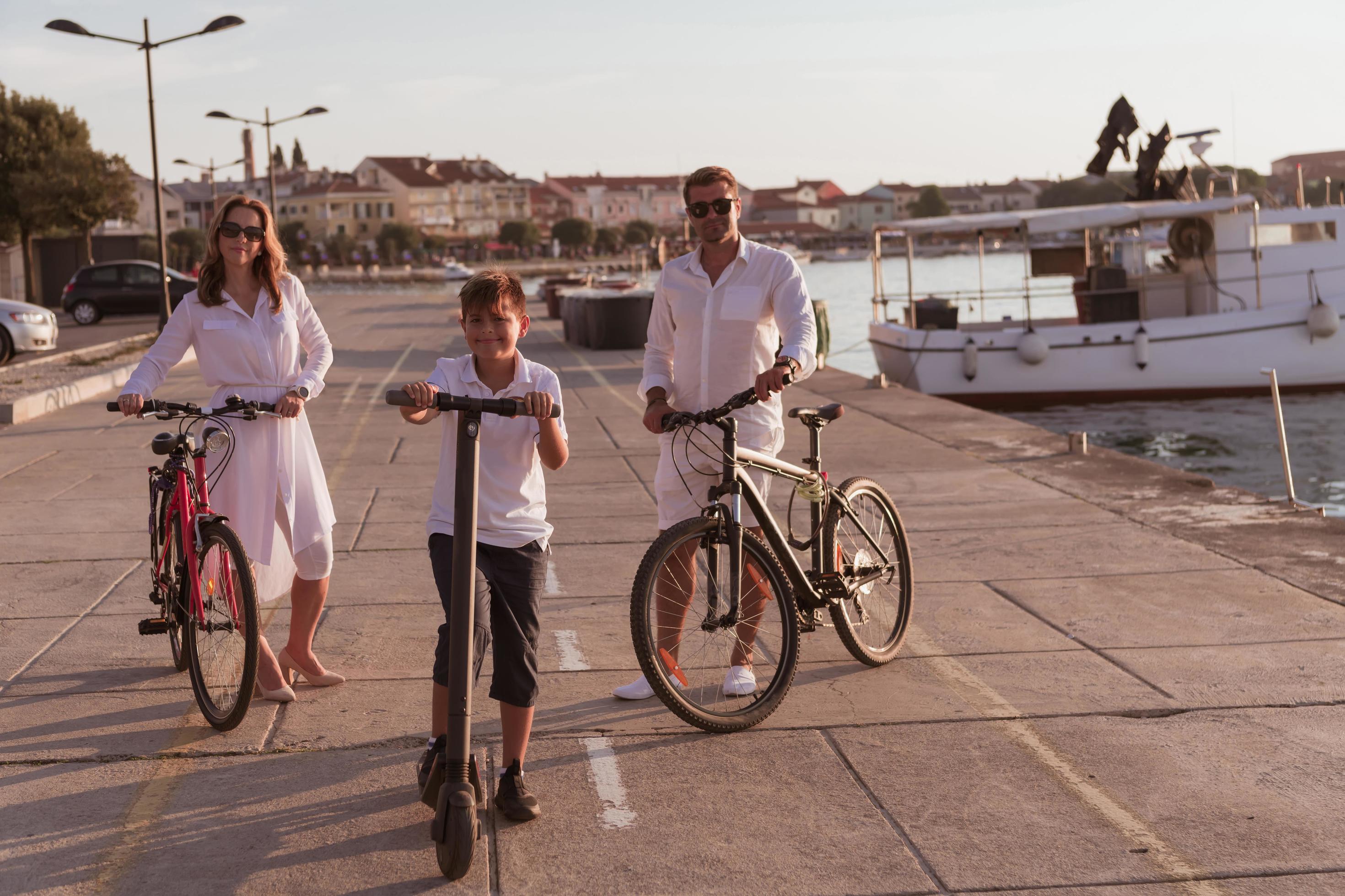 Happy family enjoying a beautiful morning by the sea together, parents riding a bike and their son riding an electric scooter. Selective focus Stock Free