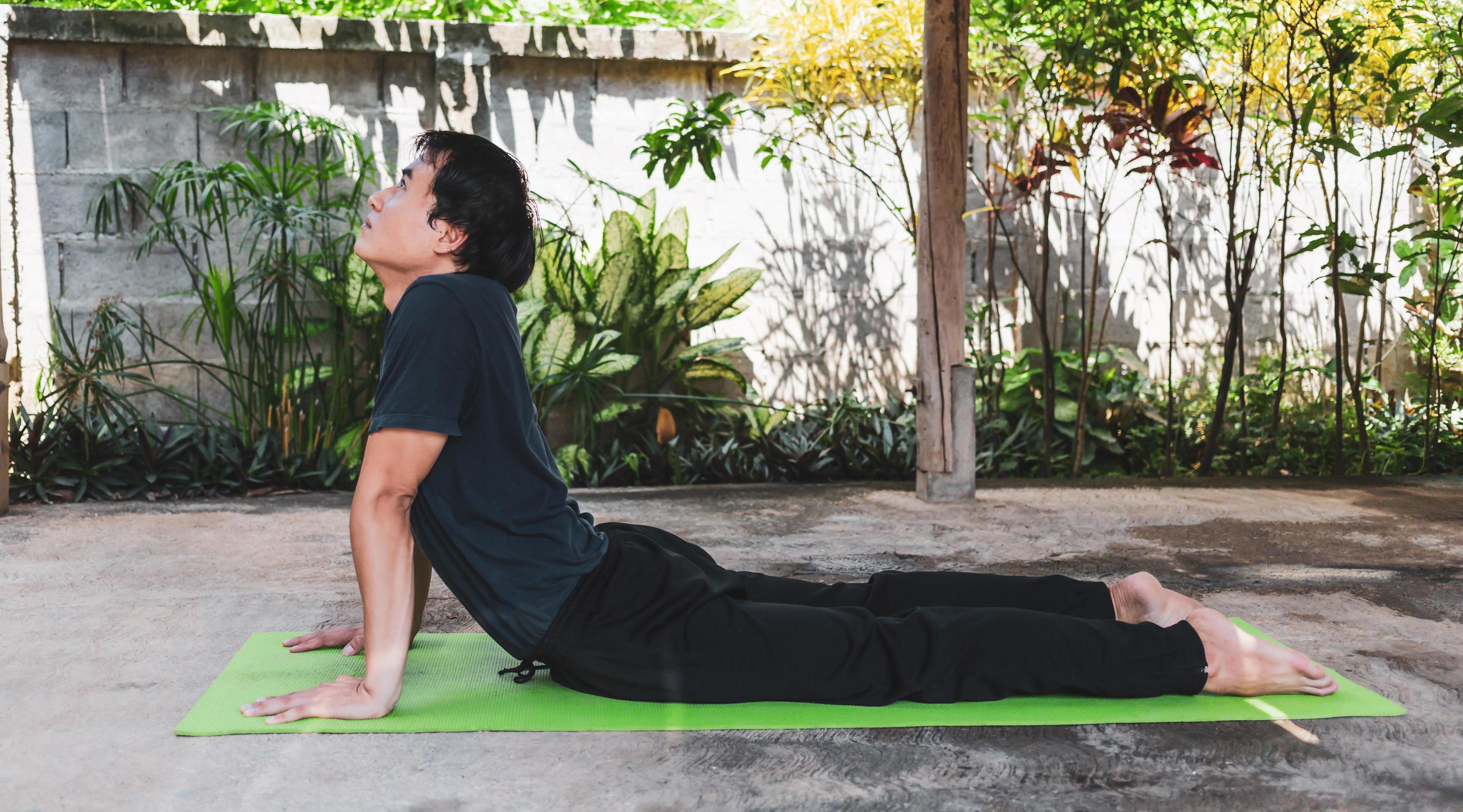 Asian man in casual outfit practicing yoga in cobra pose on the green yoga mat in the garden at home. healthy lifestyle and healthy living concept. Stock Free