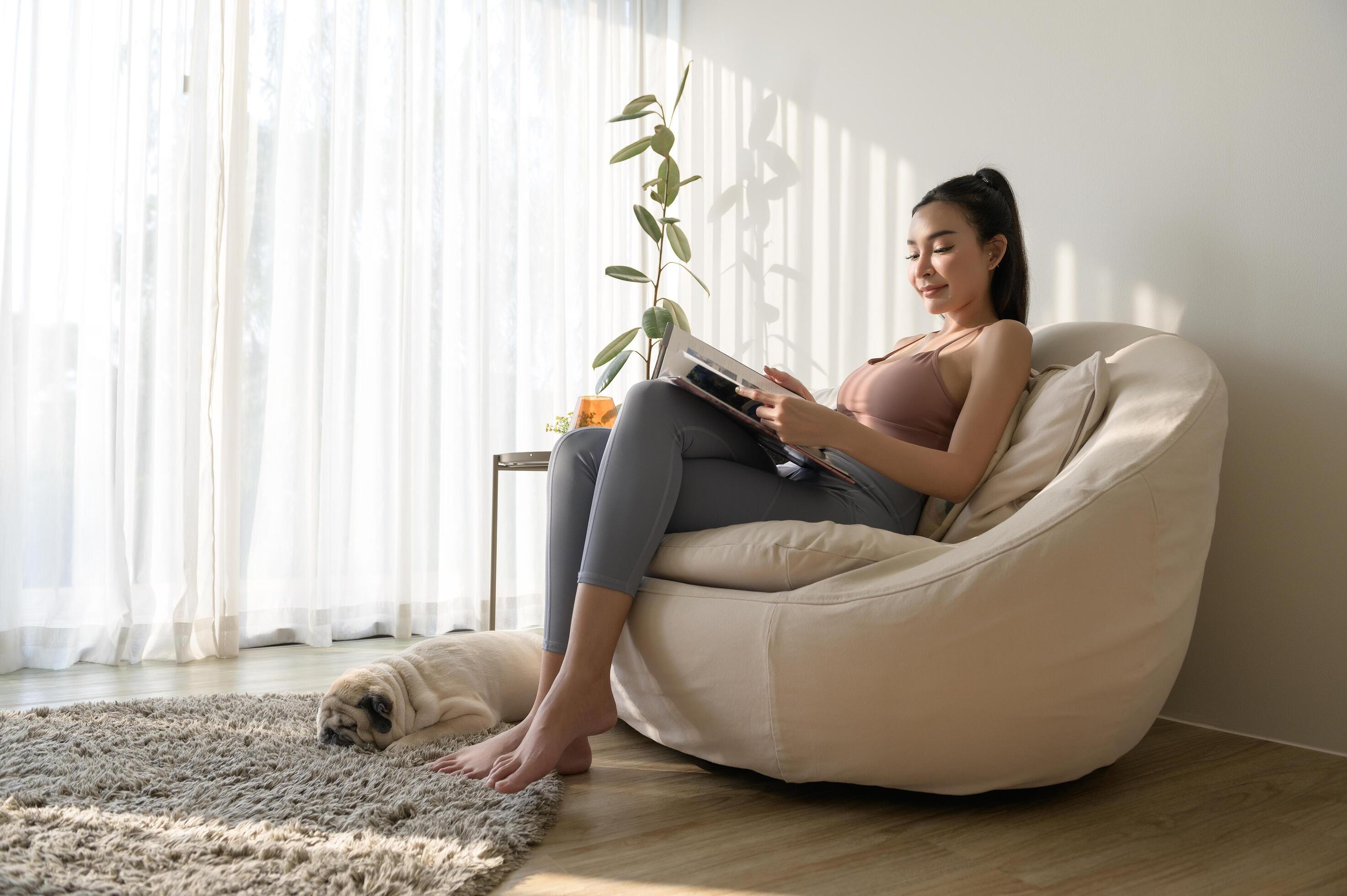 Young asian woman reading book with pug dog in living room at home Stock Free