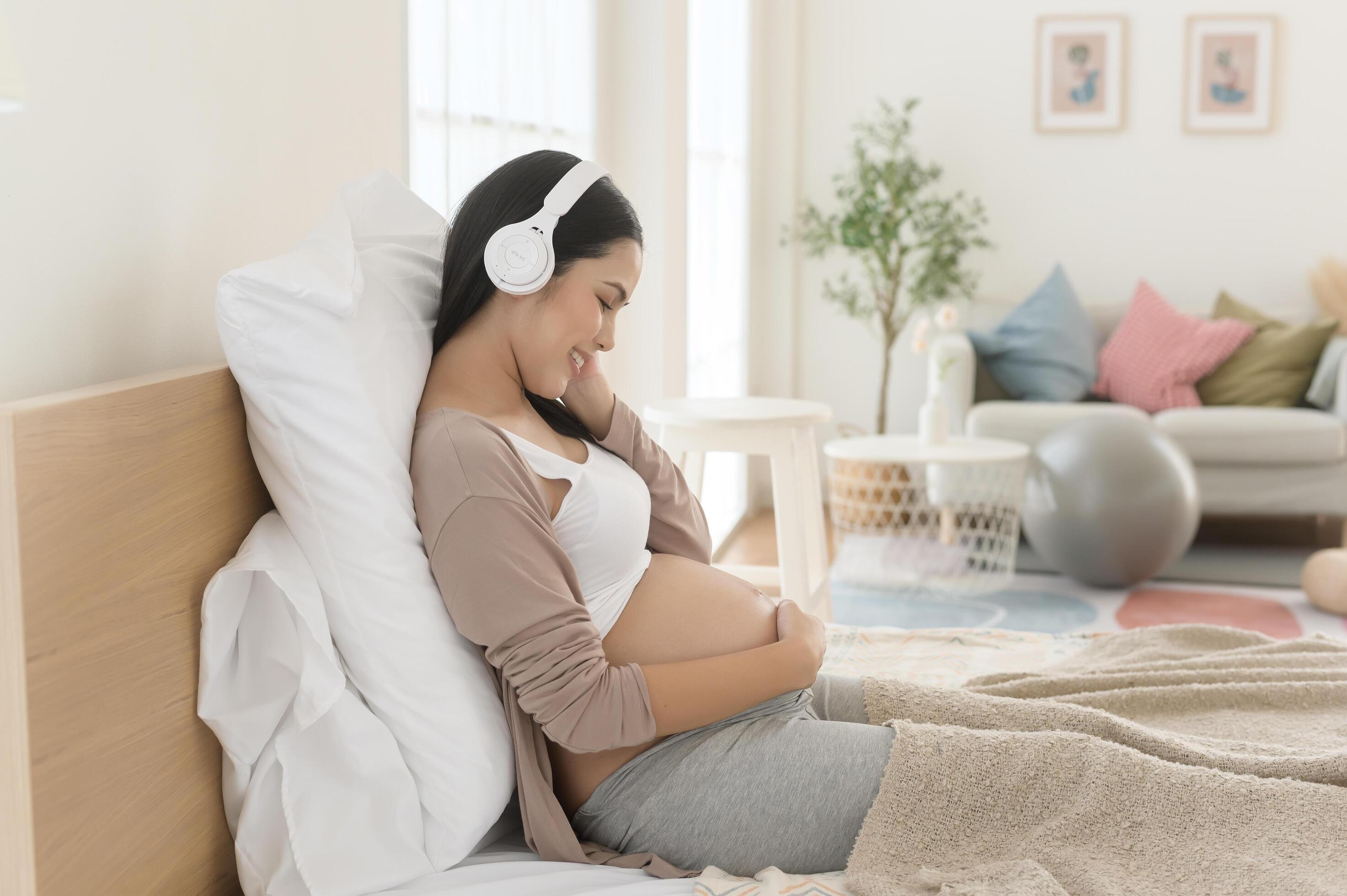 Happy pregnant woman with headphones listening to mozart music and lying on bed, pregnancy concept Stock Free