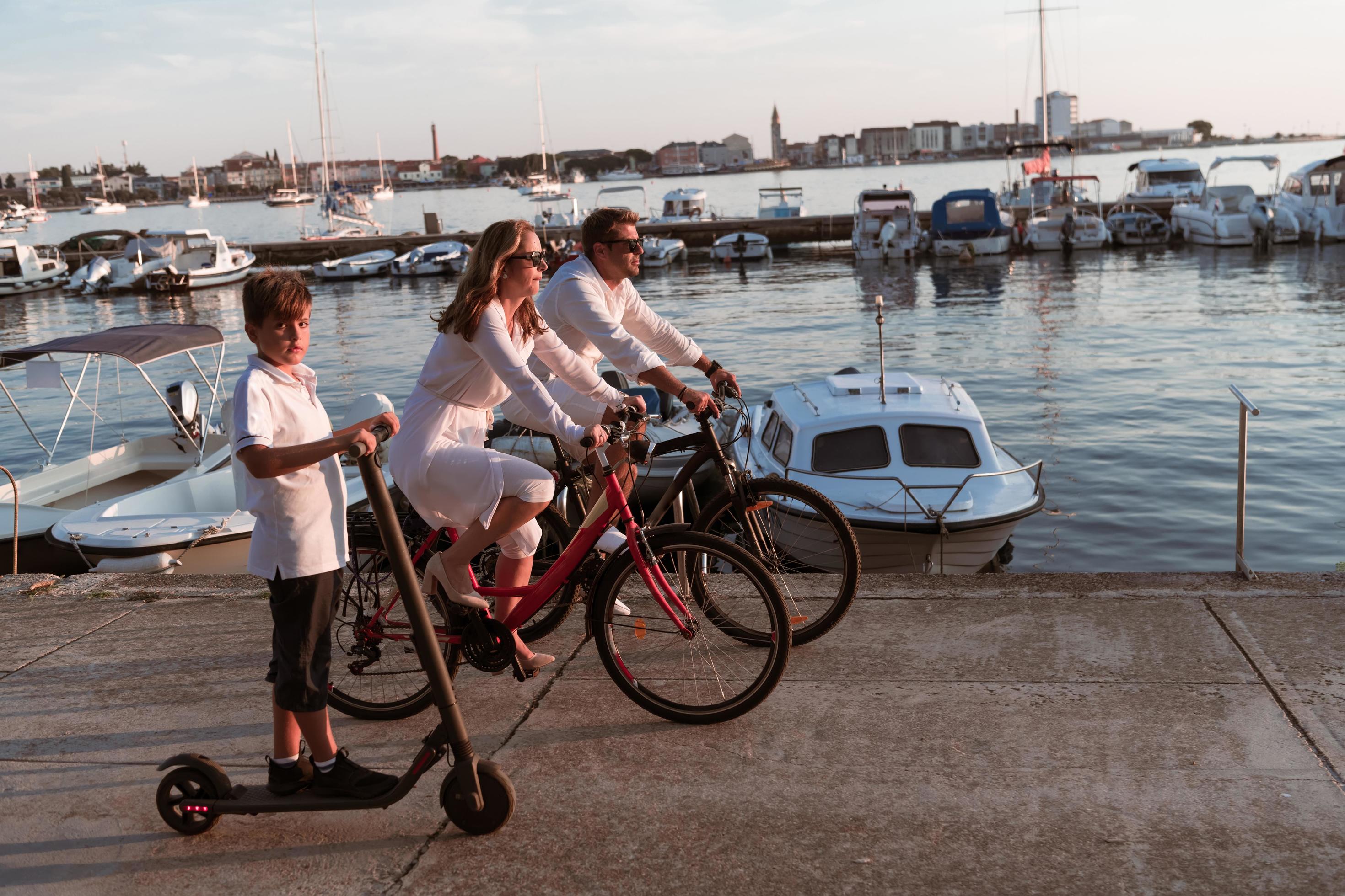 Happy family enjoying a beautiful morning by the sea together, parents riding a bike and their son riding an electric scooter. Selective focus Stock Free