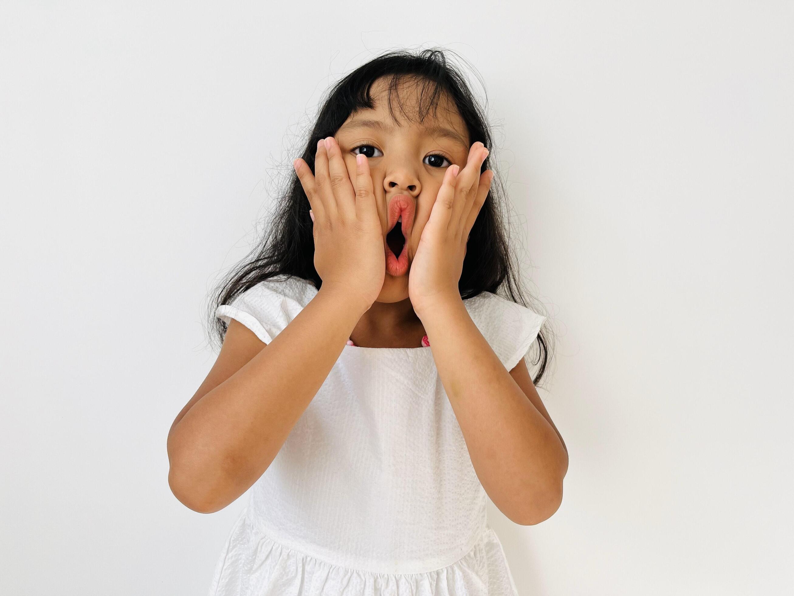 A five-year-old girl with wavy, shoulder-length black hair wearing a white dress makes a cute and silly face with her two hands pressing her lips Stock Free