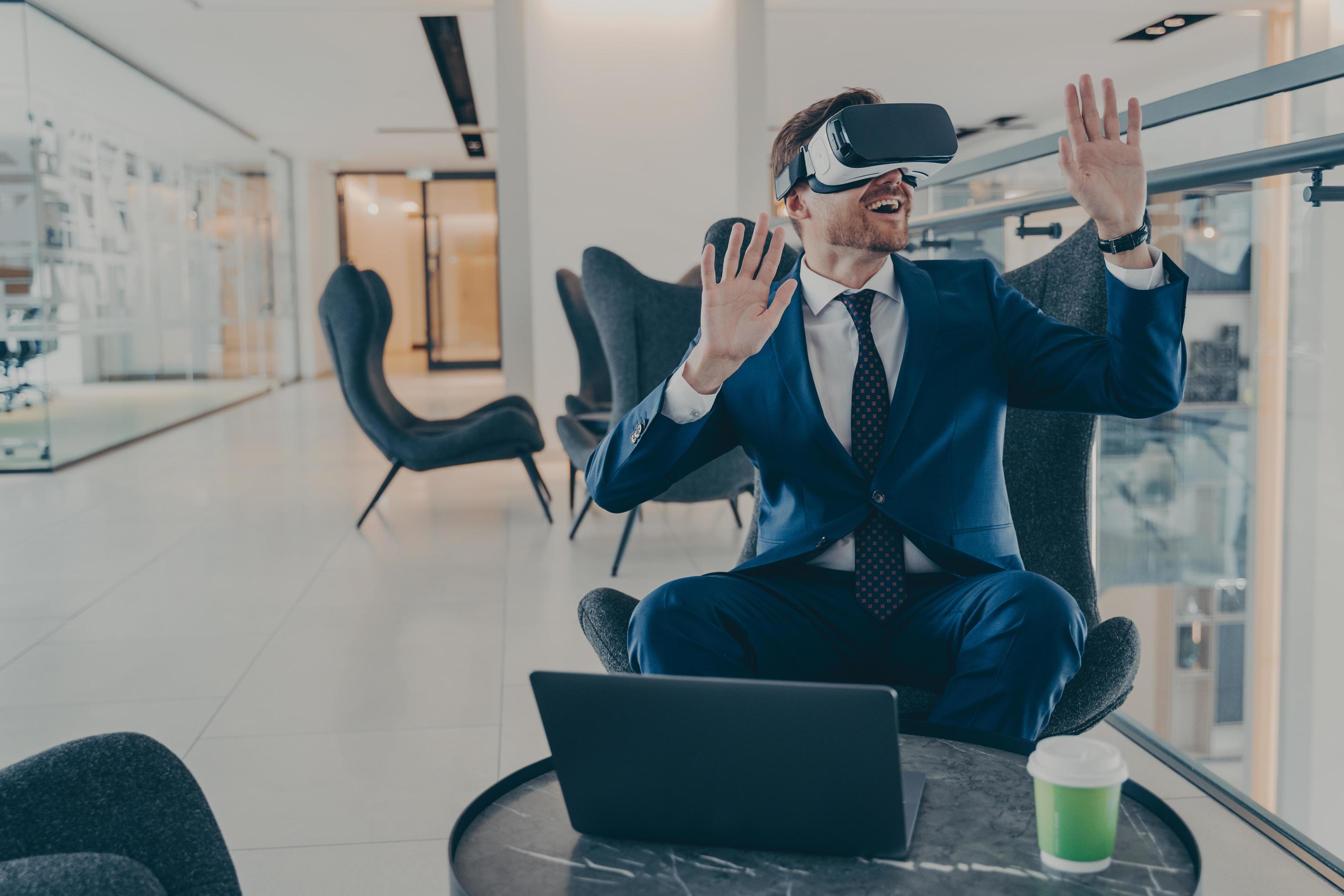 Exited CEO in VR headset enthusiastically testing app on laptop sitting in business centre lobby Stock Free