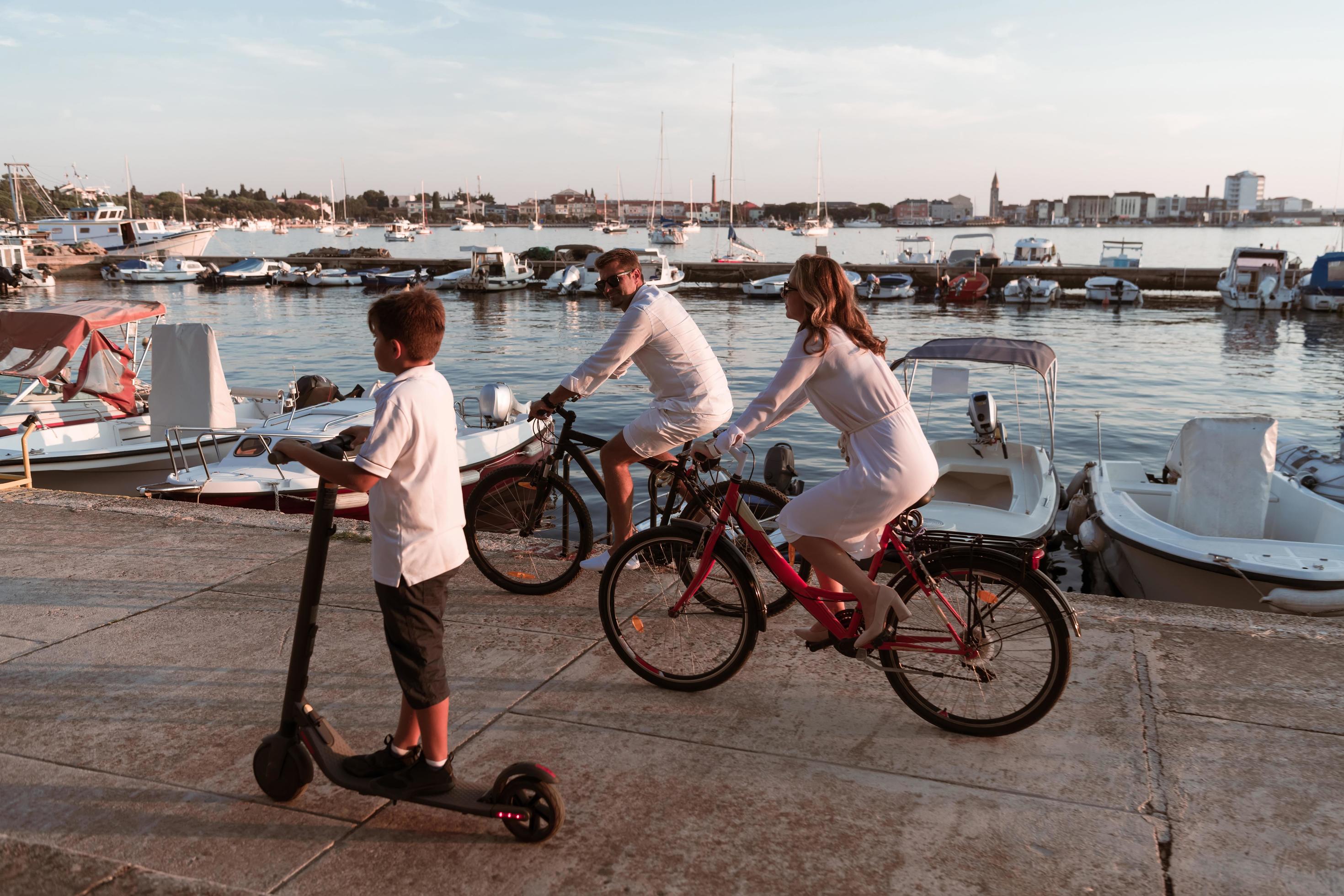 Happy family enjoying a beautiful morning by the sea together, parents riding a bike and their son riding an electric scooter. Selective focus Stock Free