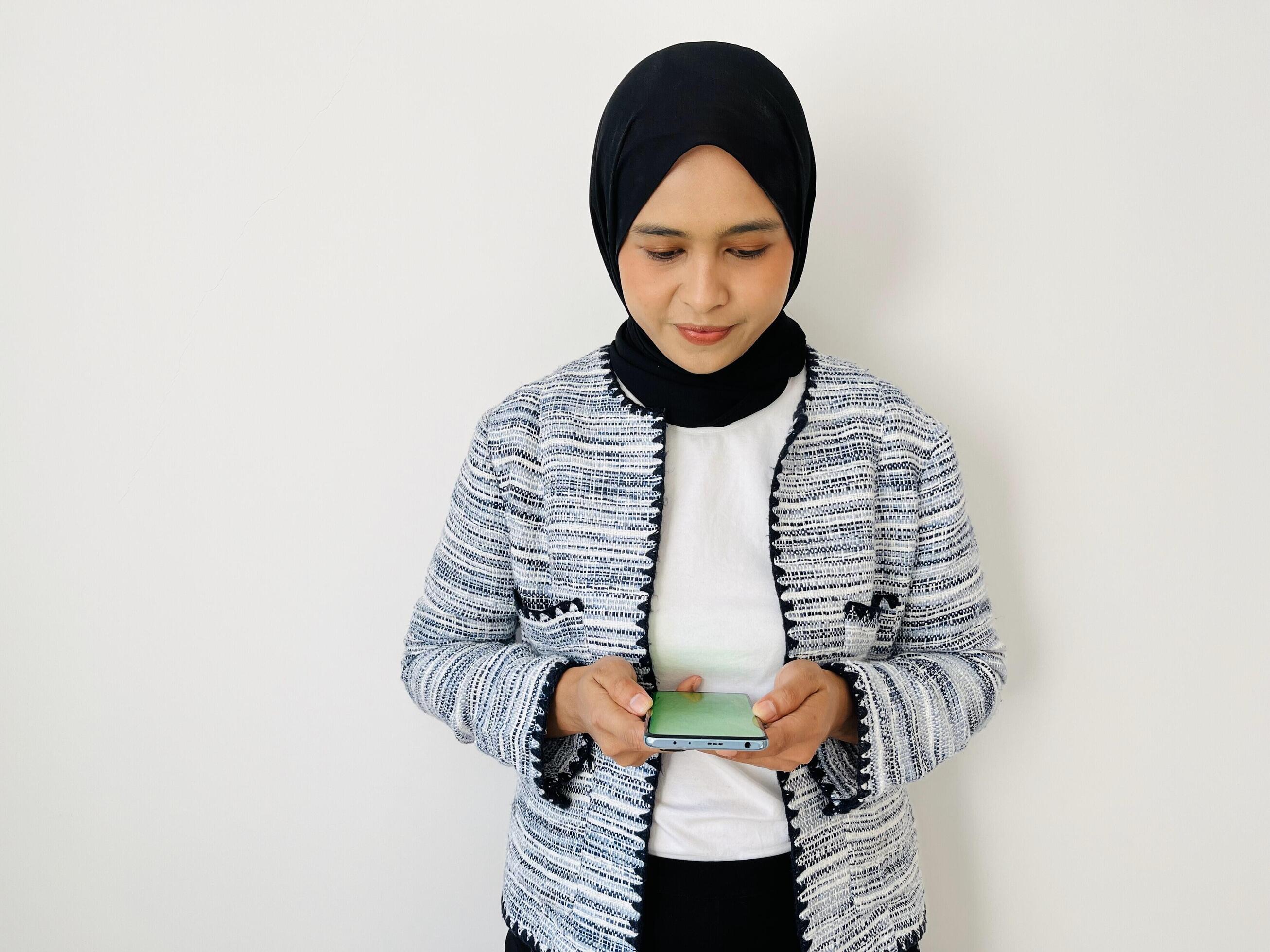 A smiling young Asian Muslim woman wear hijab and tweed blazer holding a mobile phone and looking at the mobile phone screen Stock Free