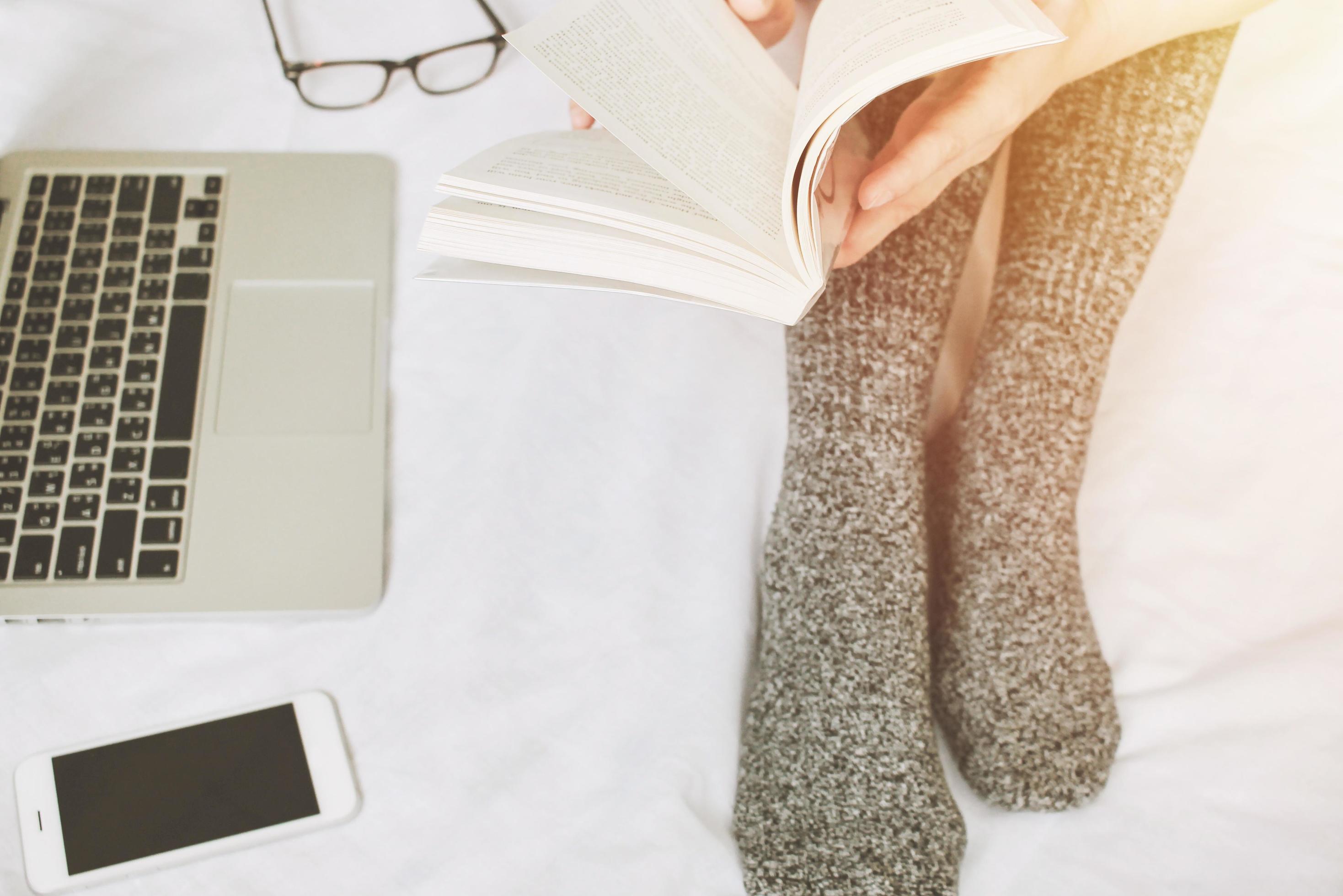 Top view of woman legs in socks reading a book and using laptop on cozy bed, lifestyle concept Stock Free