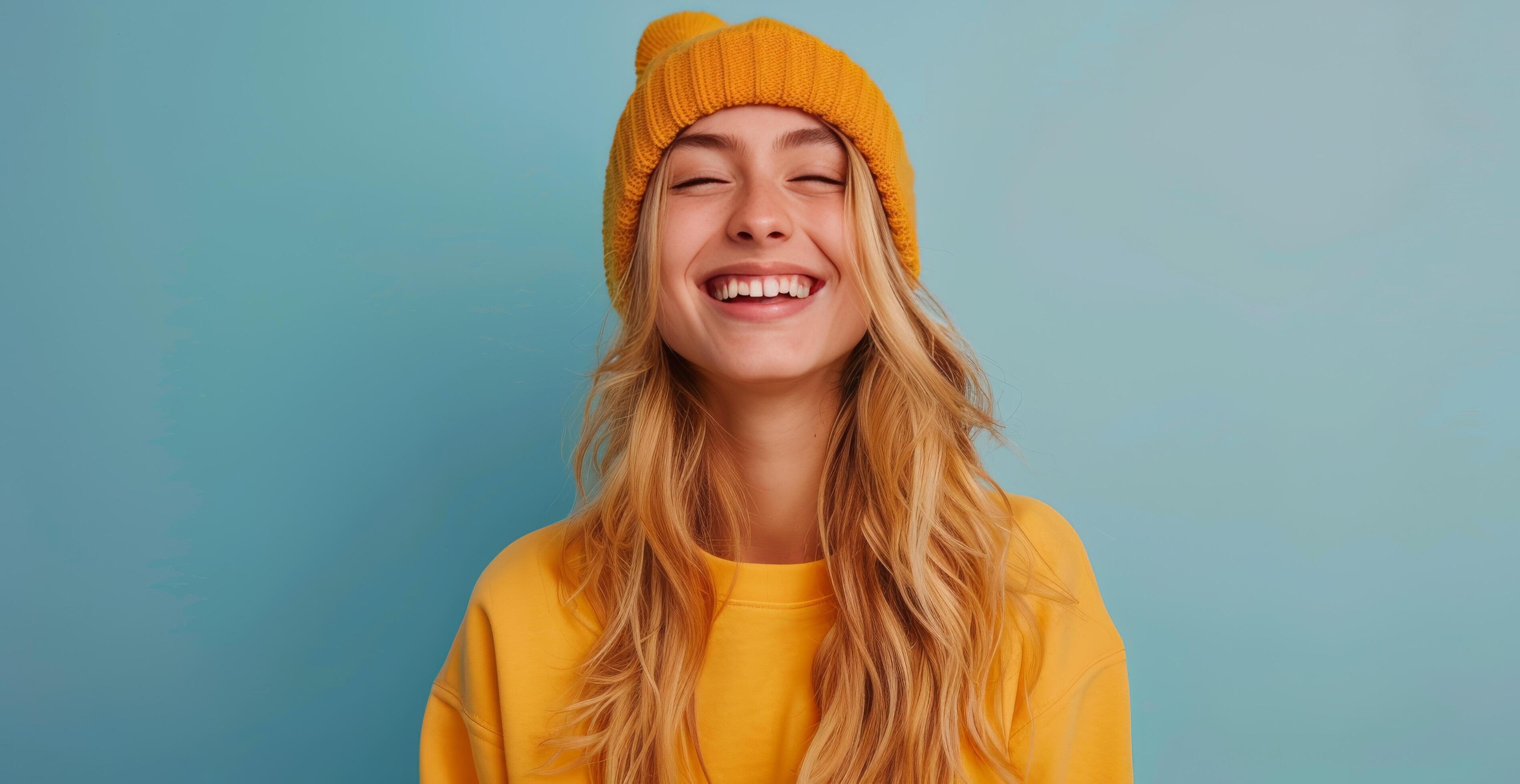 Smiling Young Woman Wearing a Yellow Hoodie and Beanie Against a Blue Background Stock Free