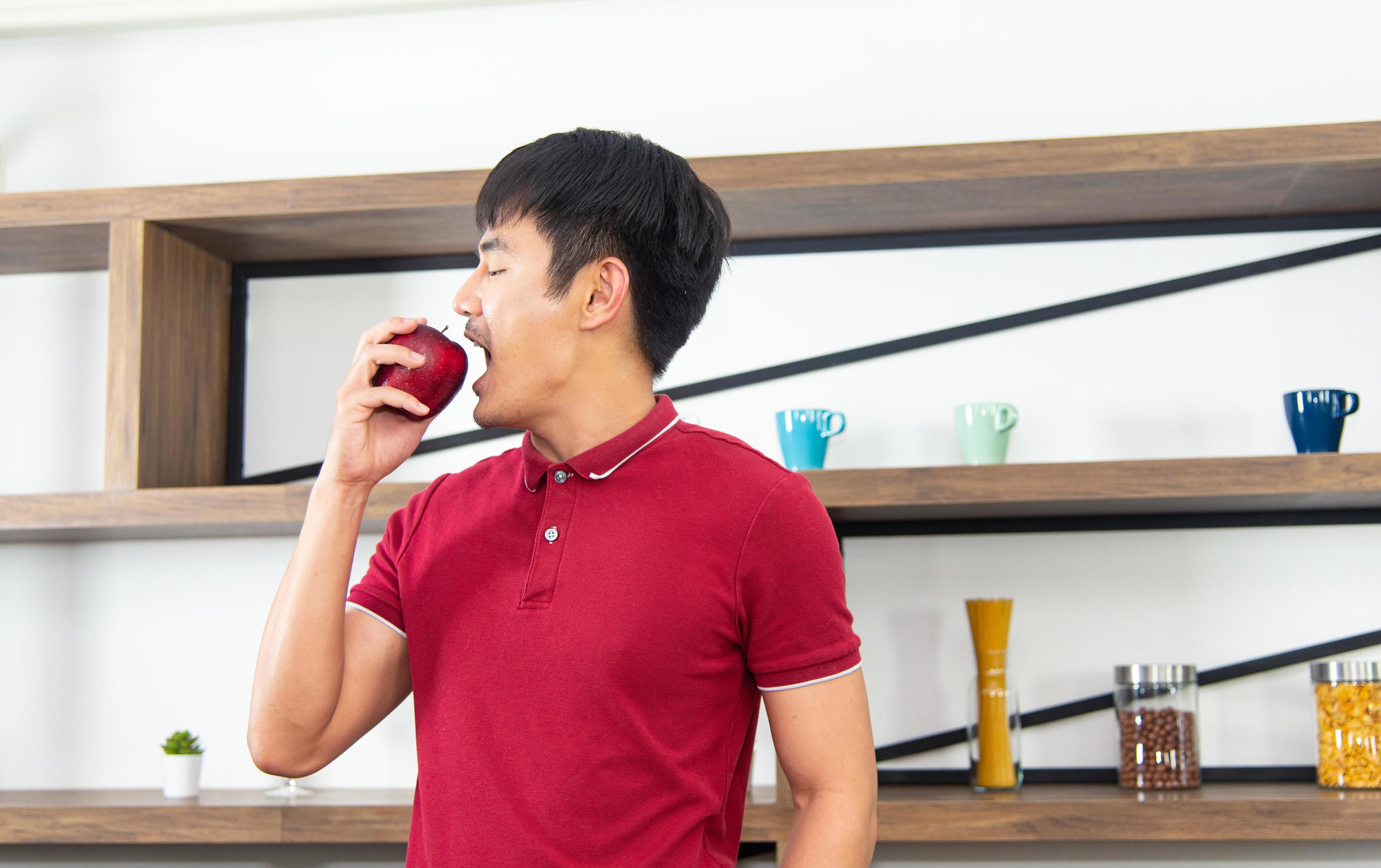 Healthy lifestyle and freshness concept. Smart, young and healthy Asian man eating apple in the loft style kitchen room. Stock Free