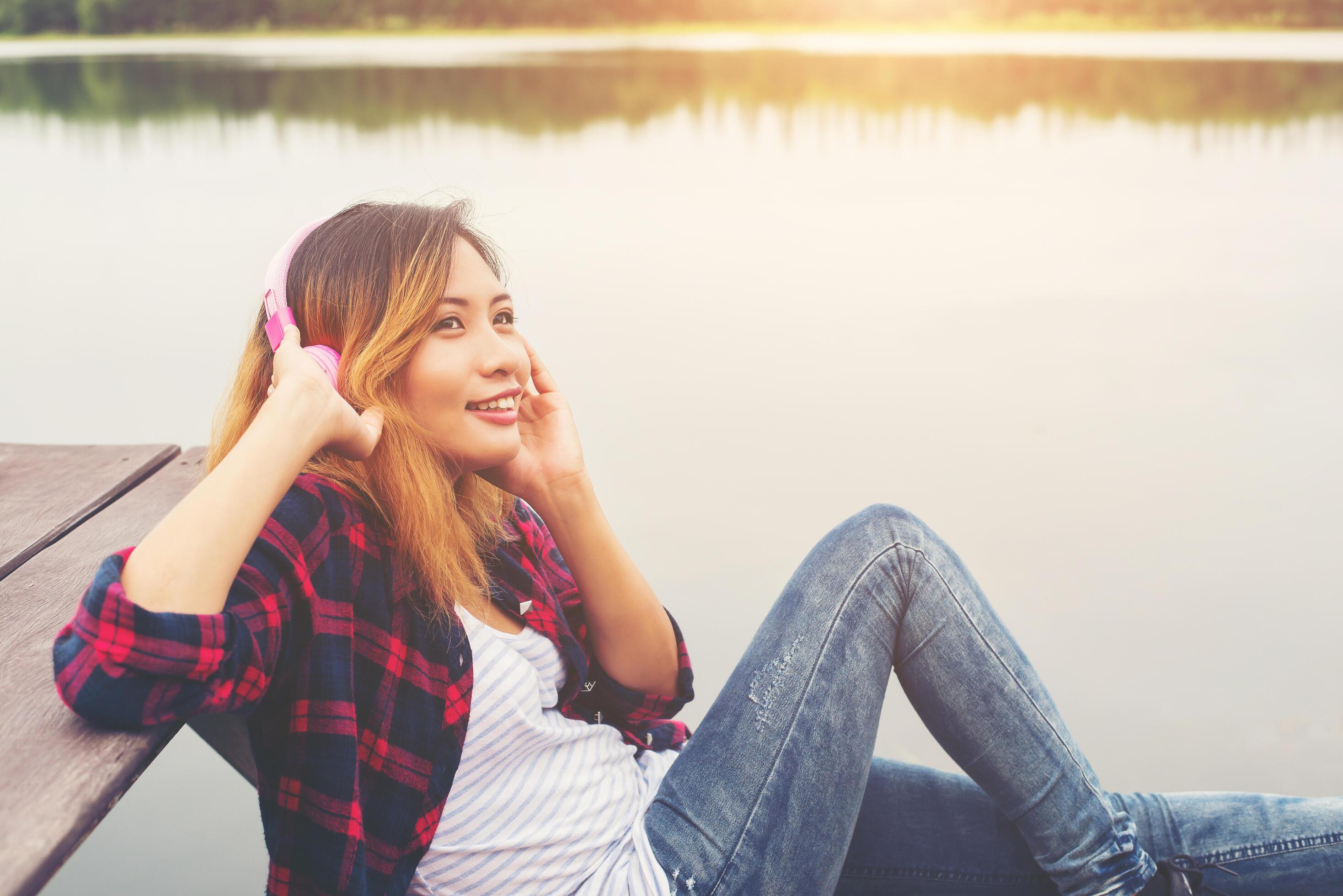 Portrait of young hipster woman with headphones sitting on pier and listening music relaxing with sunset,Lifestyle concept. Stock Free