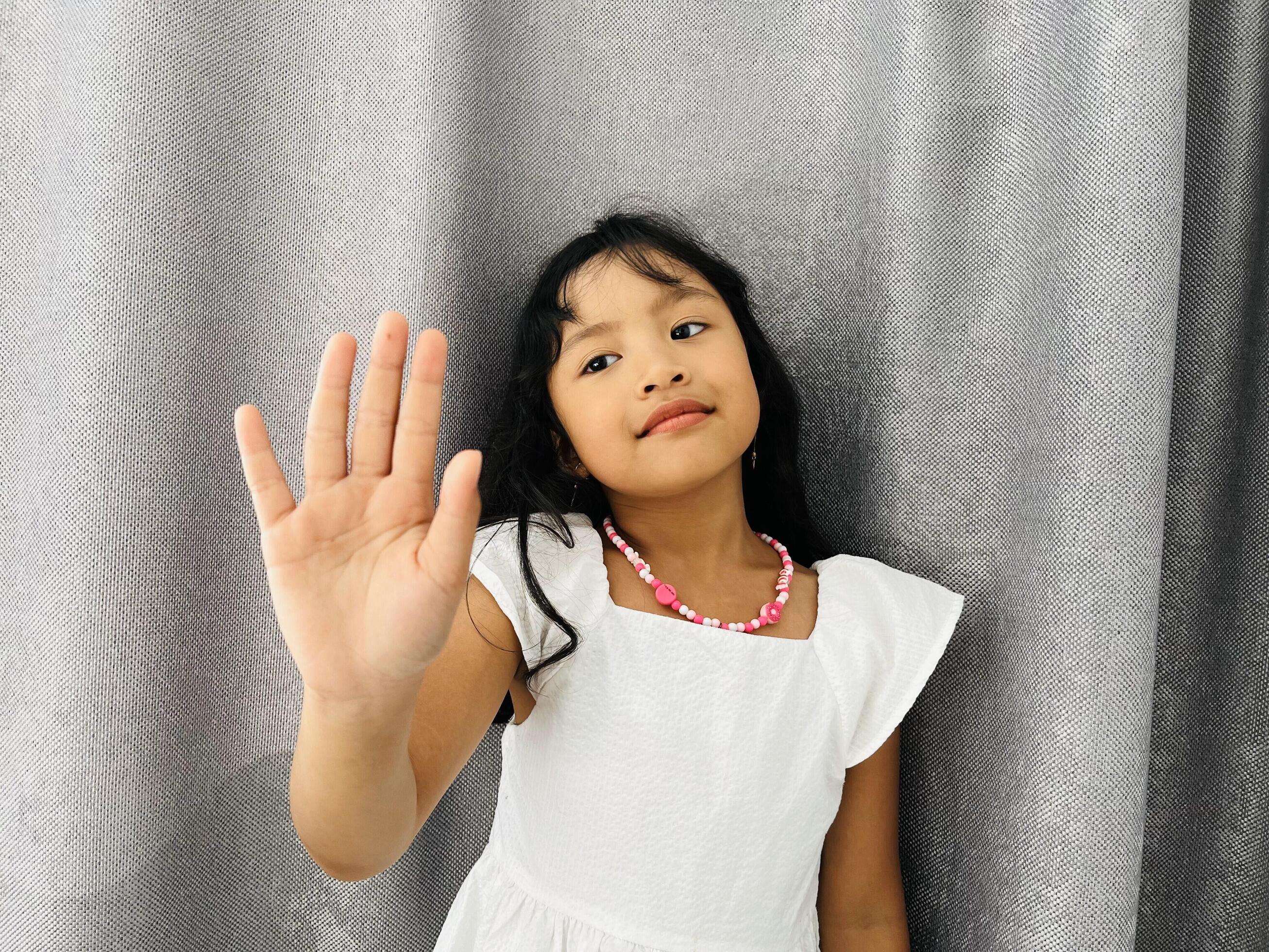 Cute adorable 5 years old girl happily shows an open hand. Little kid wearing a white dress is holding up five fingers in front of gray curtains Stock Free