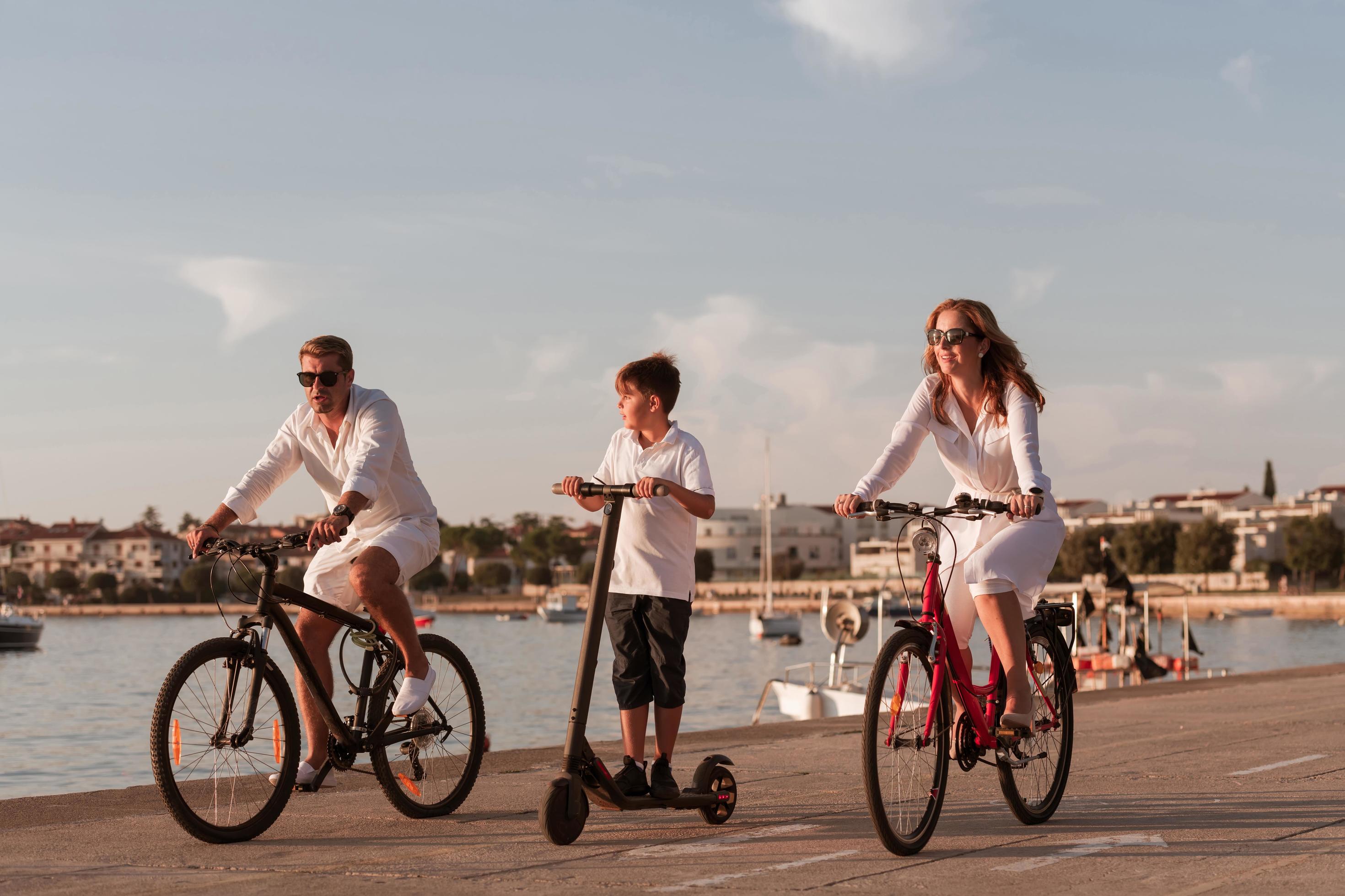 Happy family enjoying a beautiful morning by the sea together, parents riding a bike and their son riding an electric scooter. Selective focus Stock Free