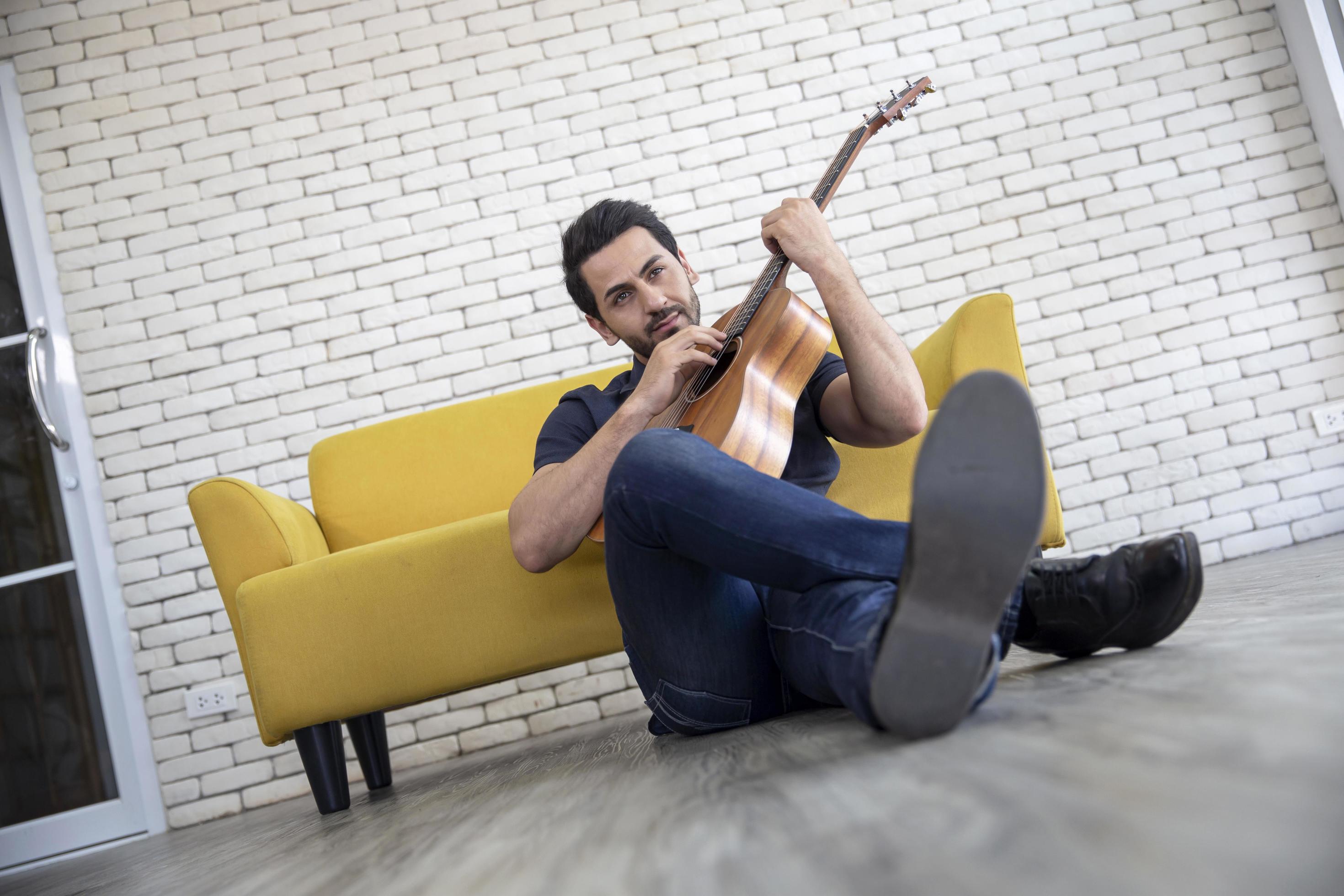 Lifestyle portrait of attractive handsome man with pleasant smile relaxing and sitting play guitar at living room Stock Free