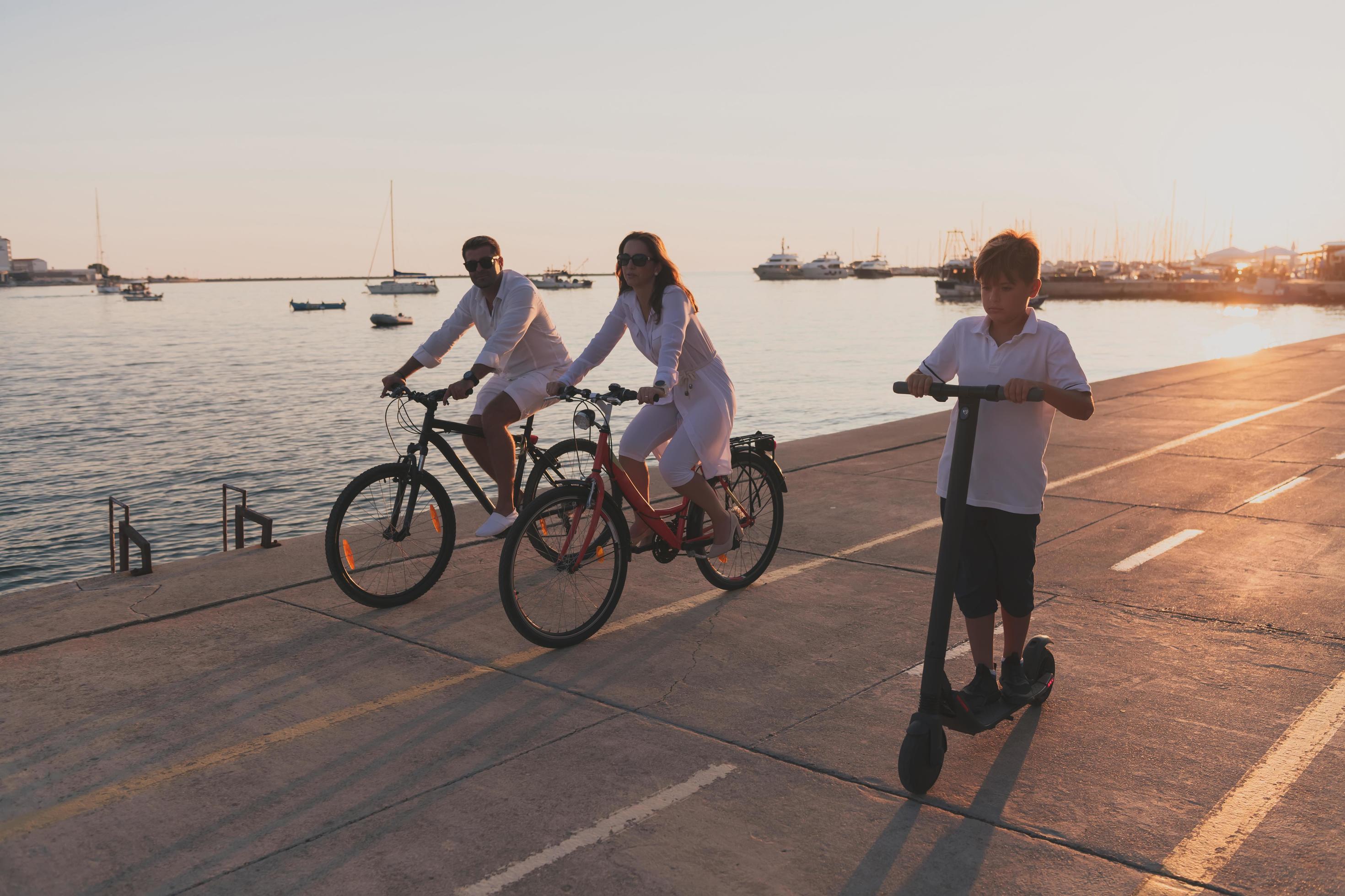 Happy family enjoying a beautiful morning by the sea together, parents riding a bike and their son riding an electric scooter. Selective focus Stock Free