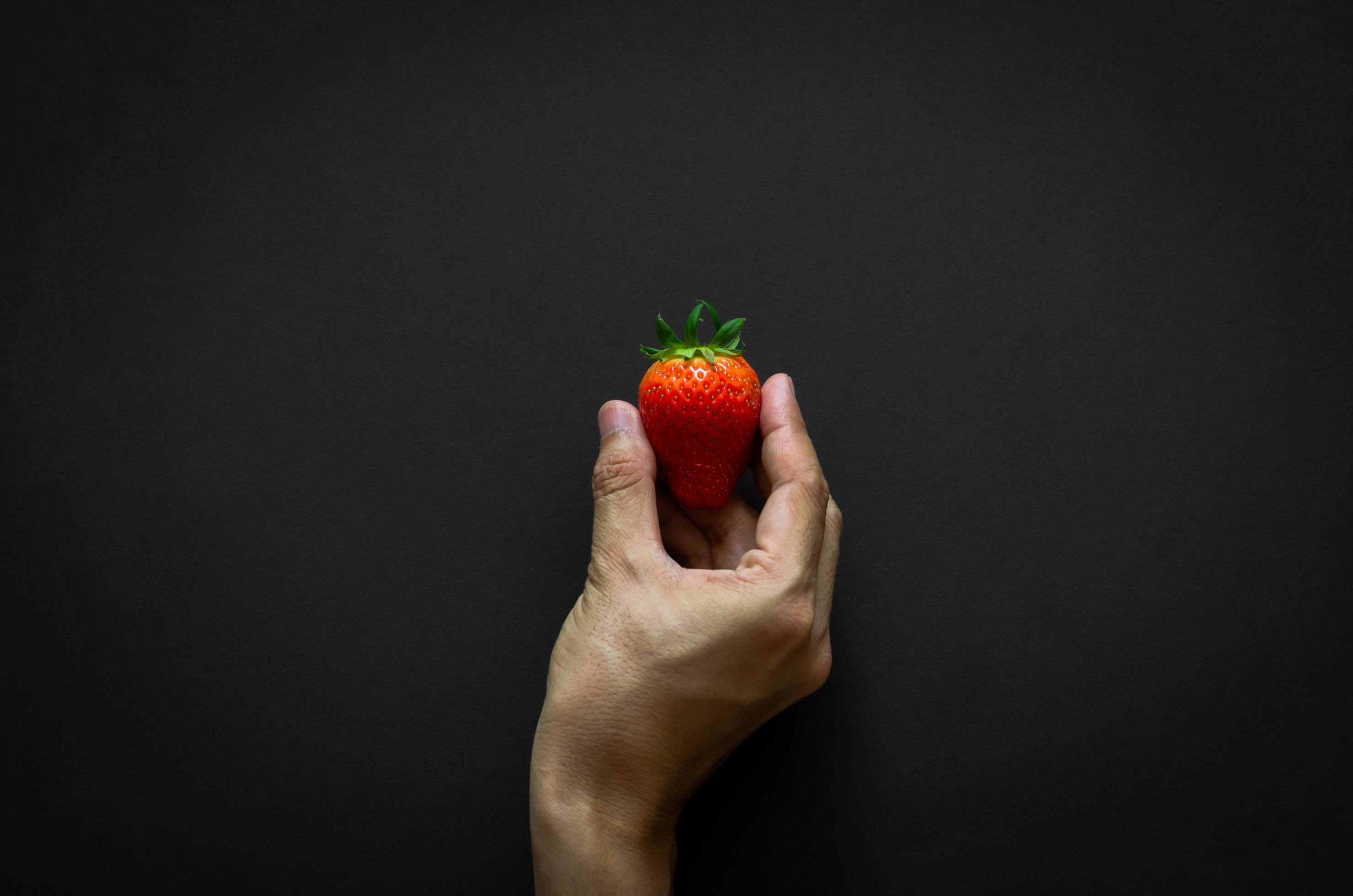 Hand holding strawberry on dark background for minimalist flat lay black food concept. Stock Free