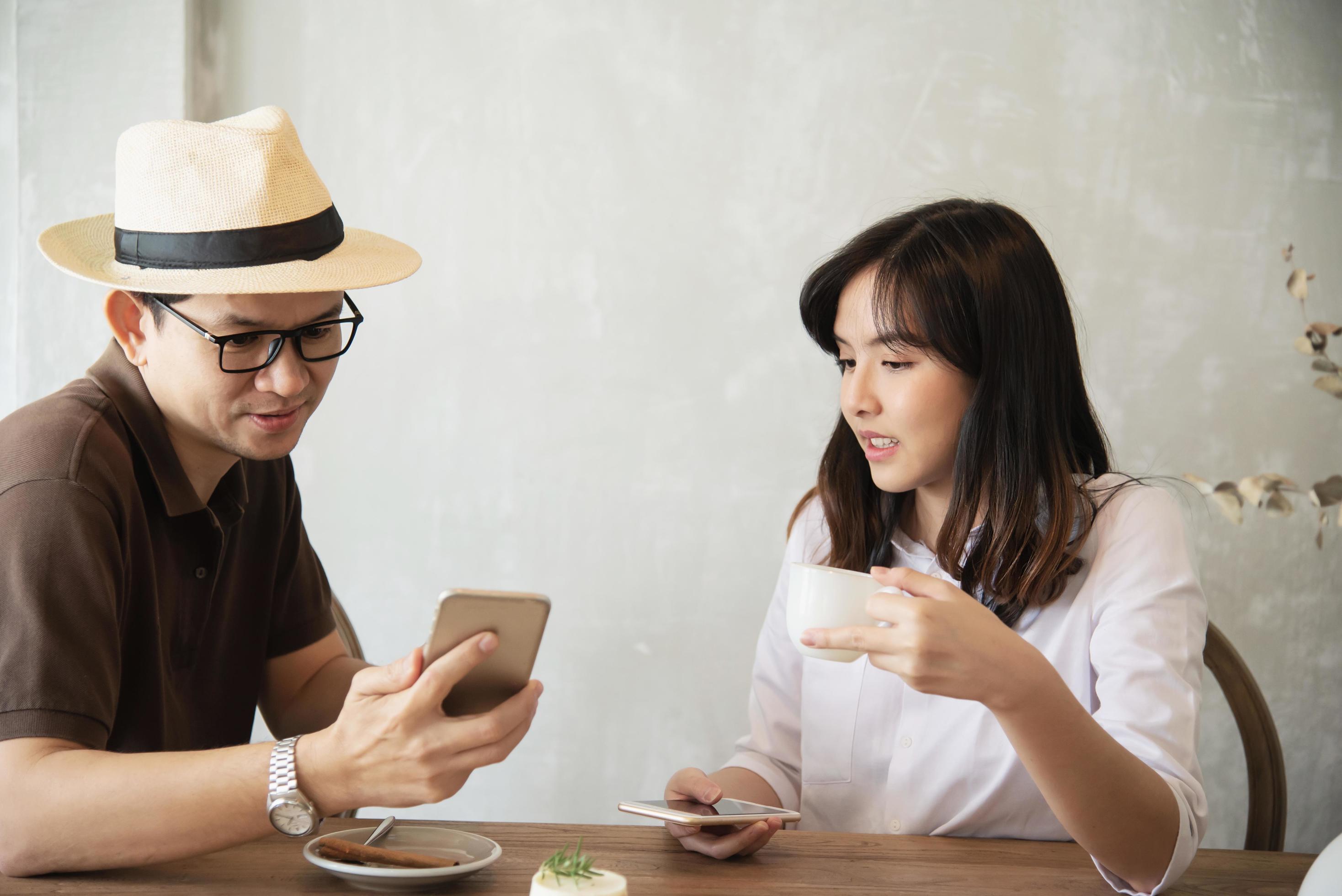 Casual man and woman talking happily while drink coffee and looking mobile phone – happy time lifestyle in coffee shop Stock Free