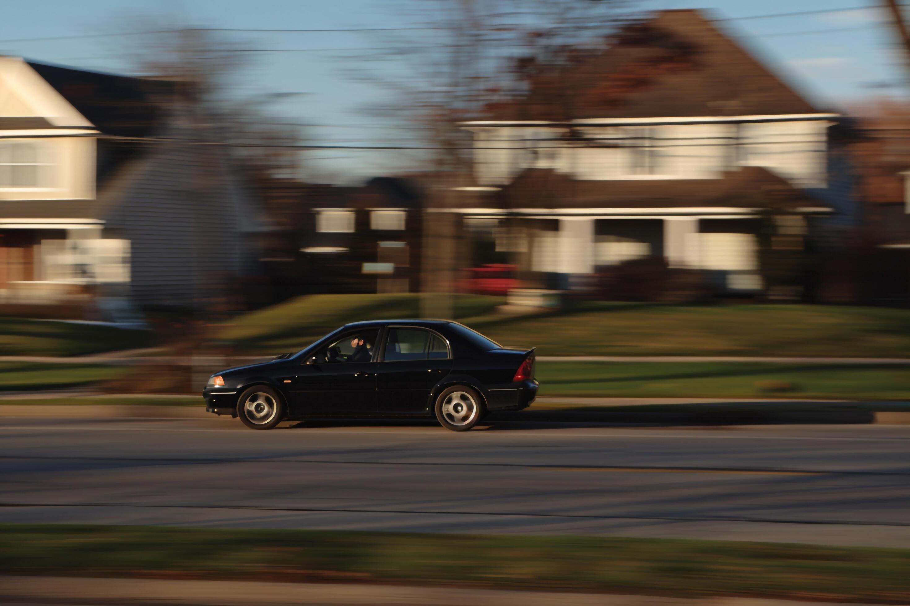 Subcompact sedan driving through suburban neighborhood with neatly trimmed lawns and family homes in the background Stock Free