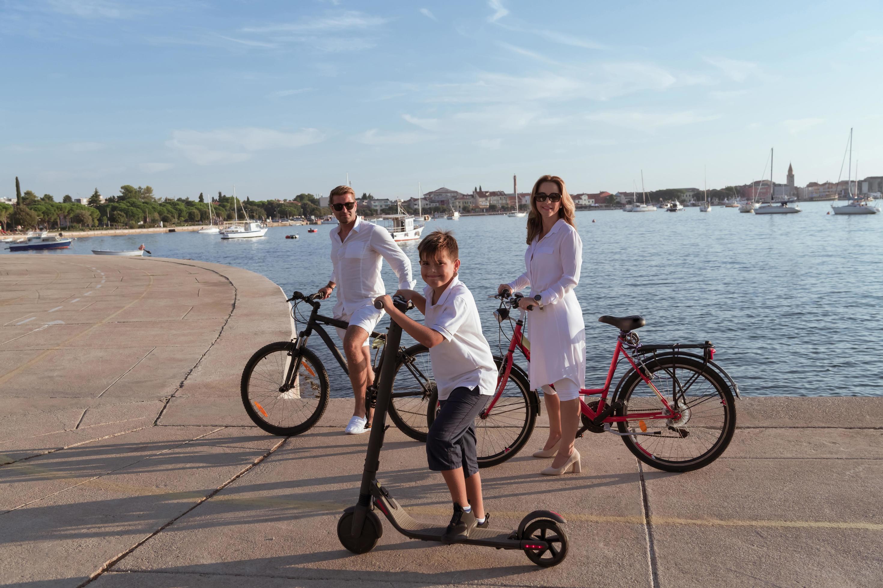 Happy family enjoying a beautiful morning by the sea together, parents riding a bike and their son riding an electric scooter. Selective focus Stock Free