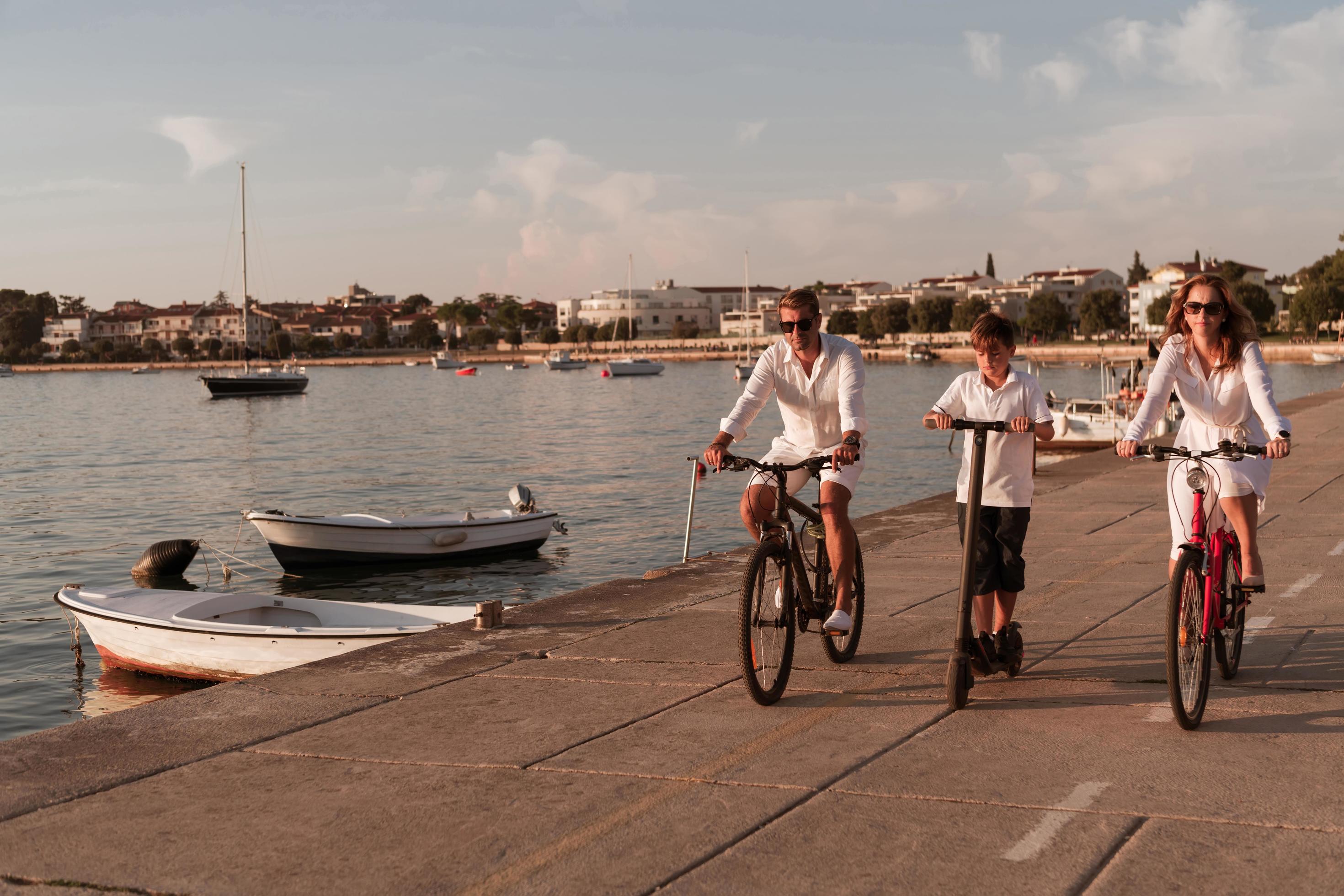 Happy family enjoying a beautiful morning by the sea together, parents riding a bike and their son riding an electric scooter. Selective focus Stock Free