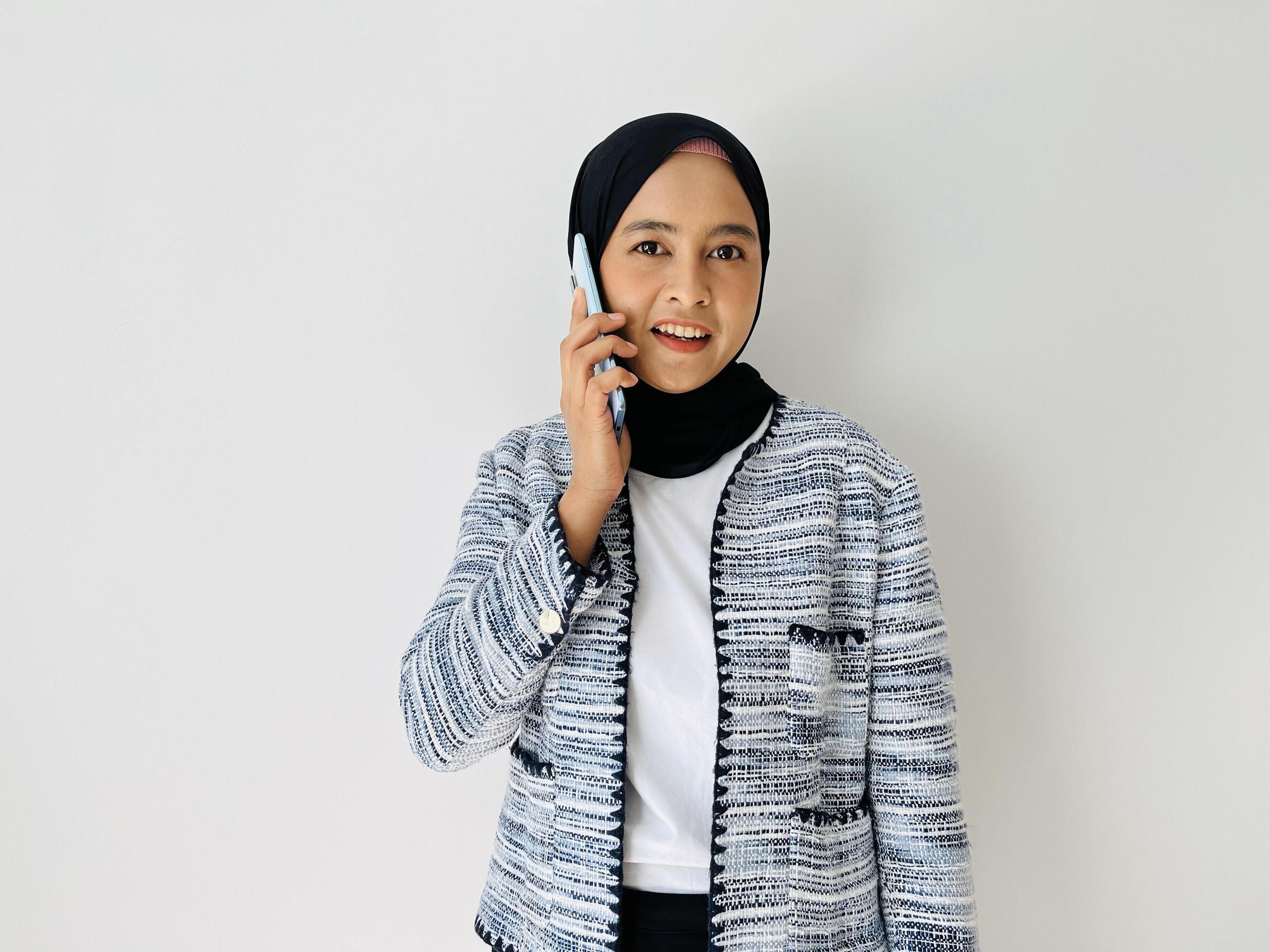 Young Asian woman smiling while using mobile phone having call. Standing female wearing black hijab and tweed blazer isolated on white background. Stock Free