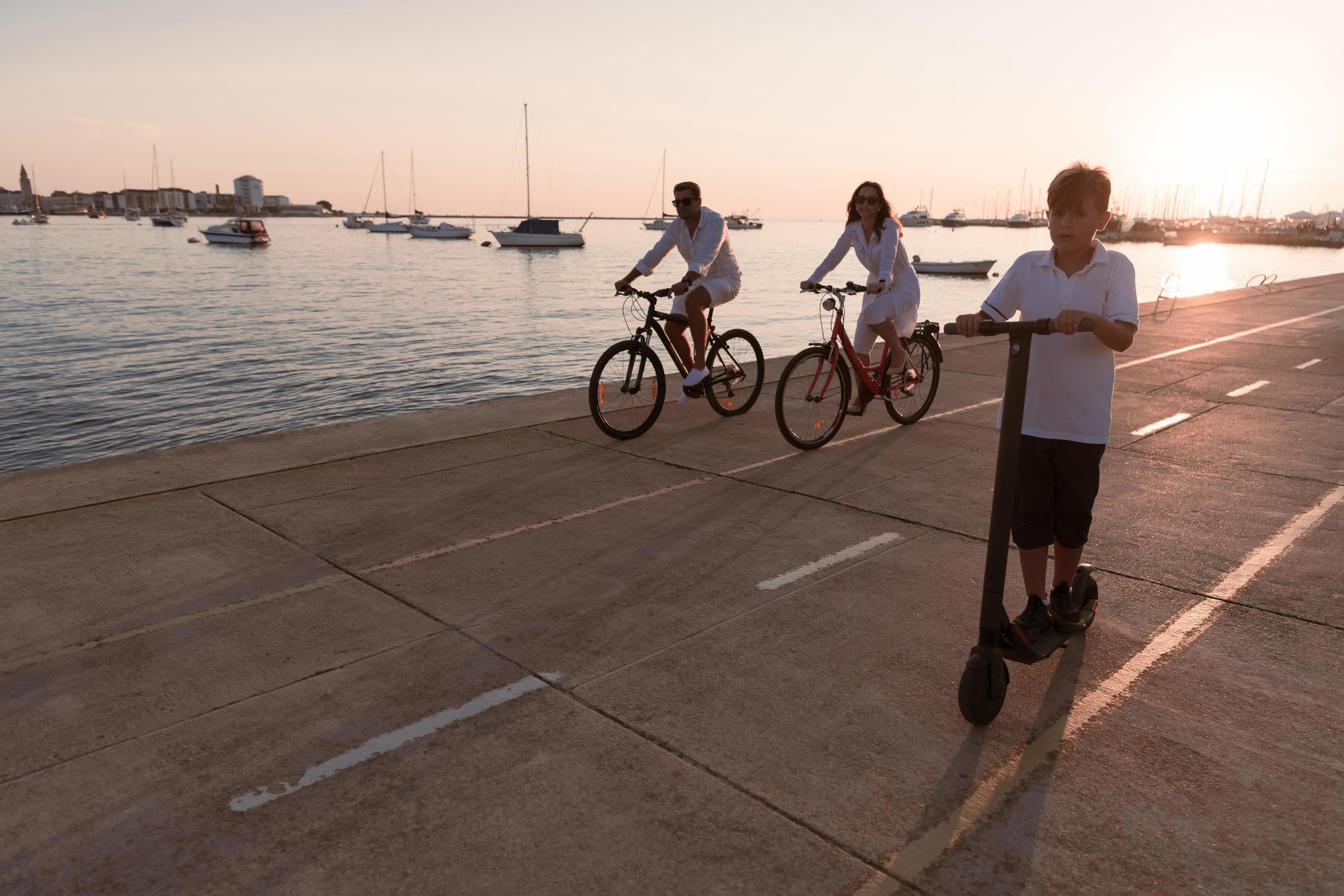 Happy family enjoying a beautiful morning by the sea together, parents riding a bike and their son riding an electric scooter. Selective focus Stock Free