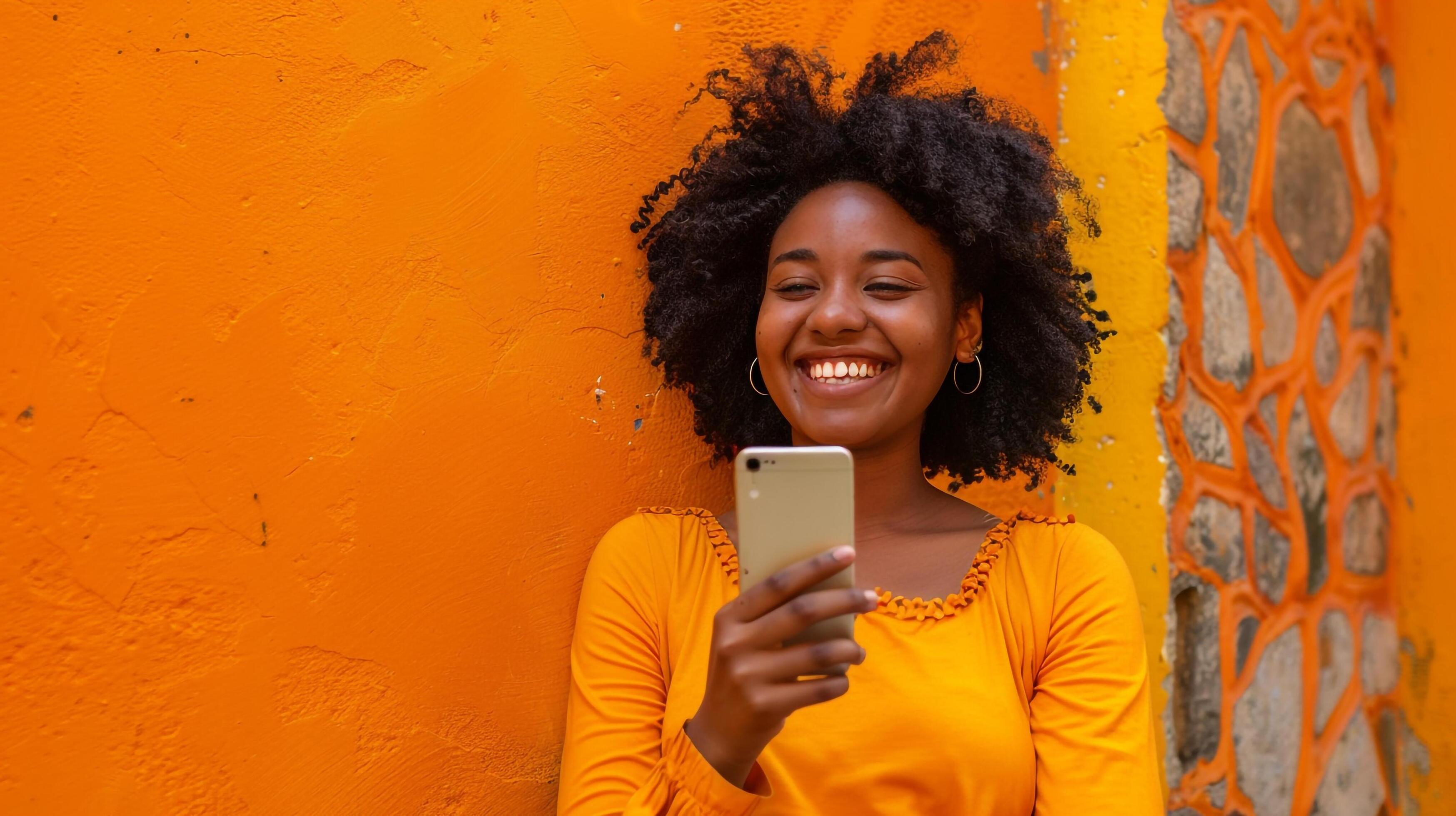 Happy woman in yellow shirt using mobile phone by wall Stock Free