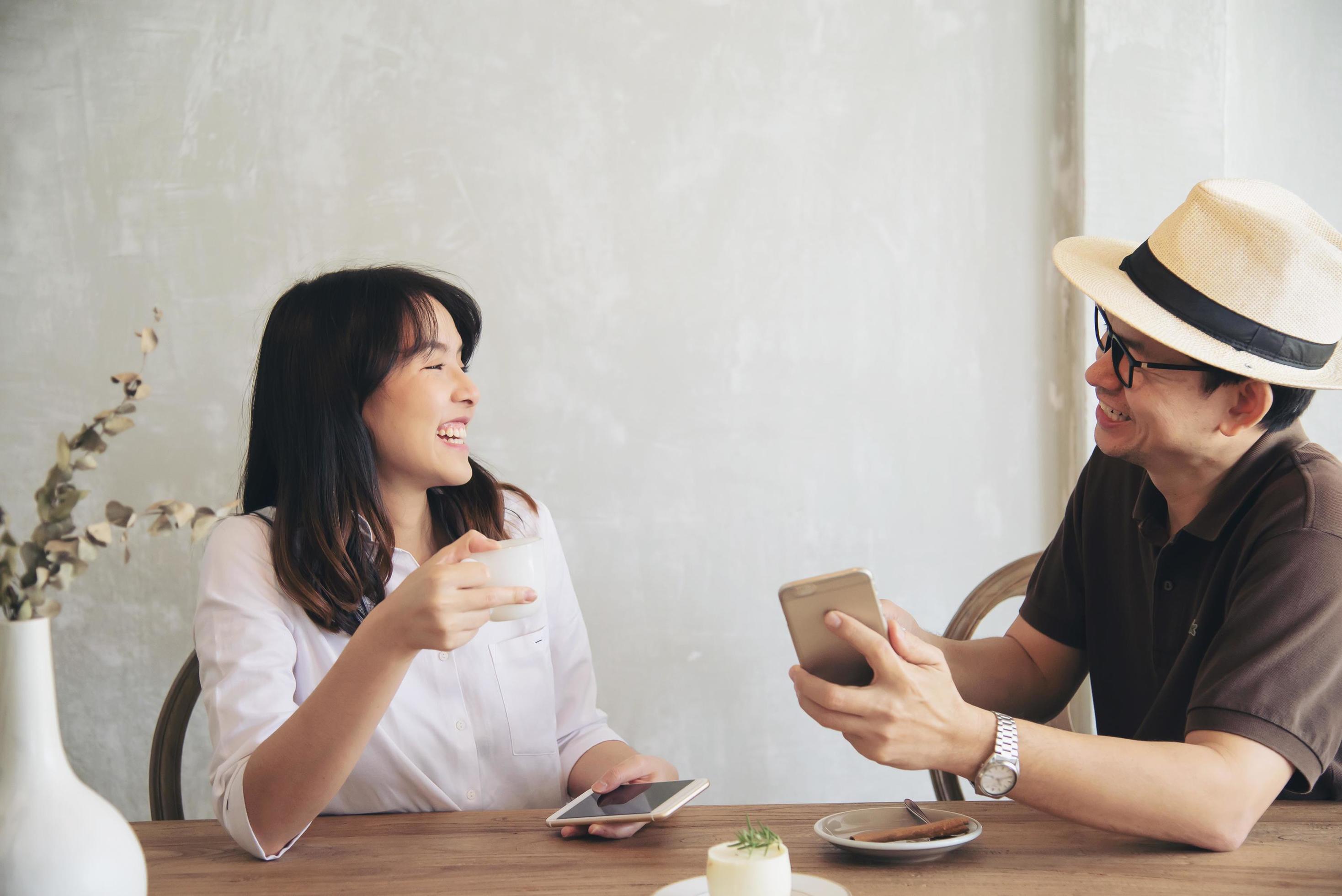 Casual man and woman talking happily while drink coffee and looking mobile phone – happy time lifestyle in coffee shop Stock Free