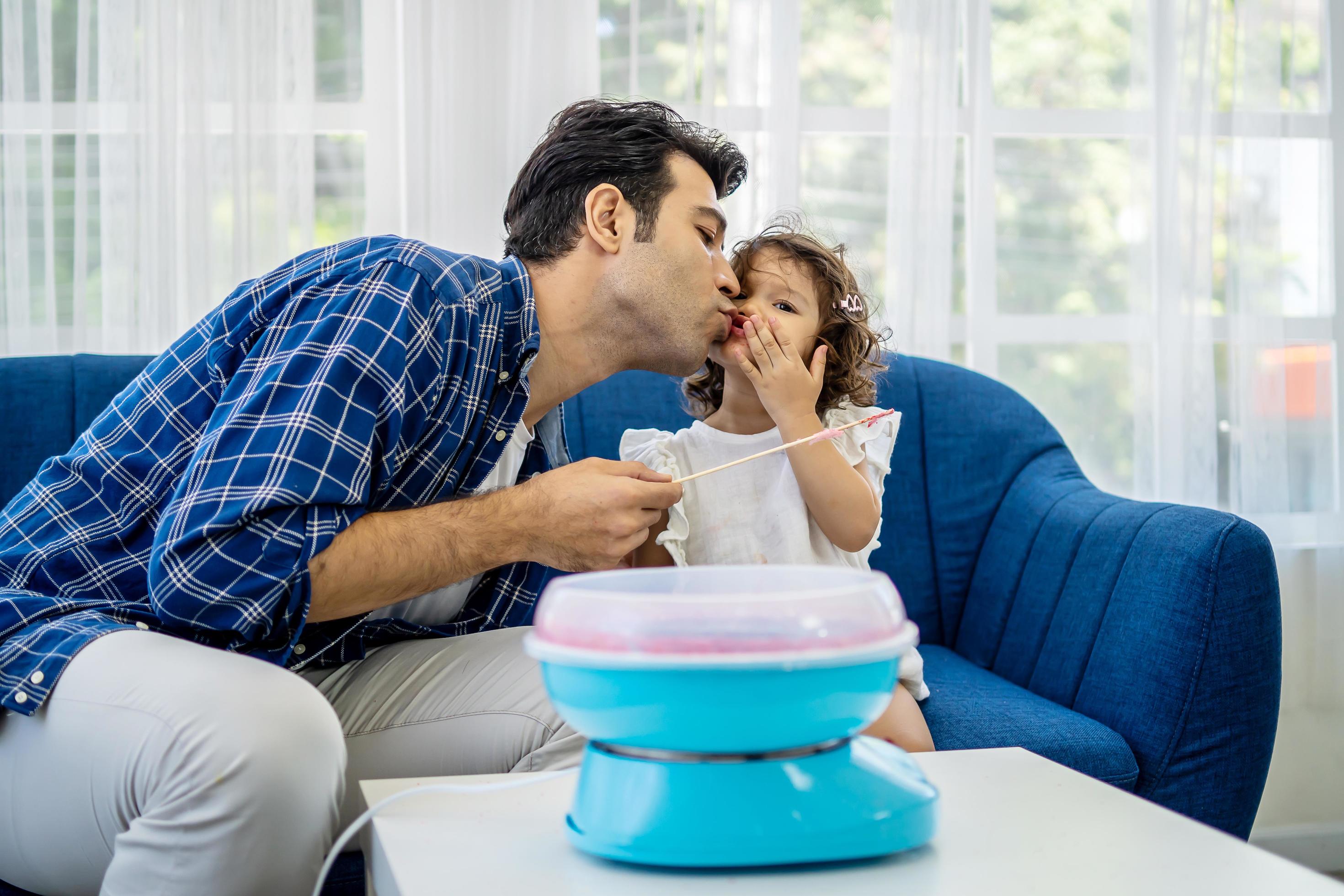 Caucasian family young father kissing his little daughter at mouth after eat cotton candy together on sofa at new house Stock Free