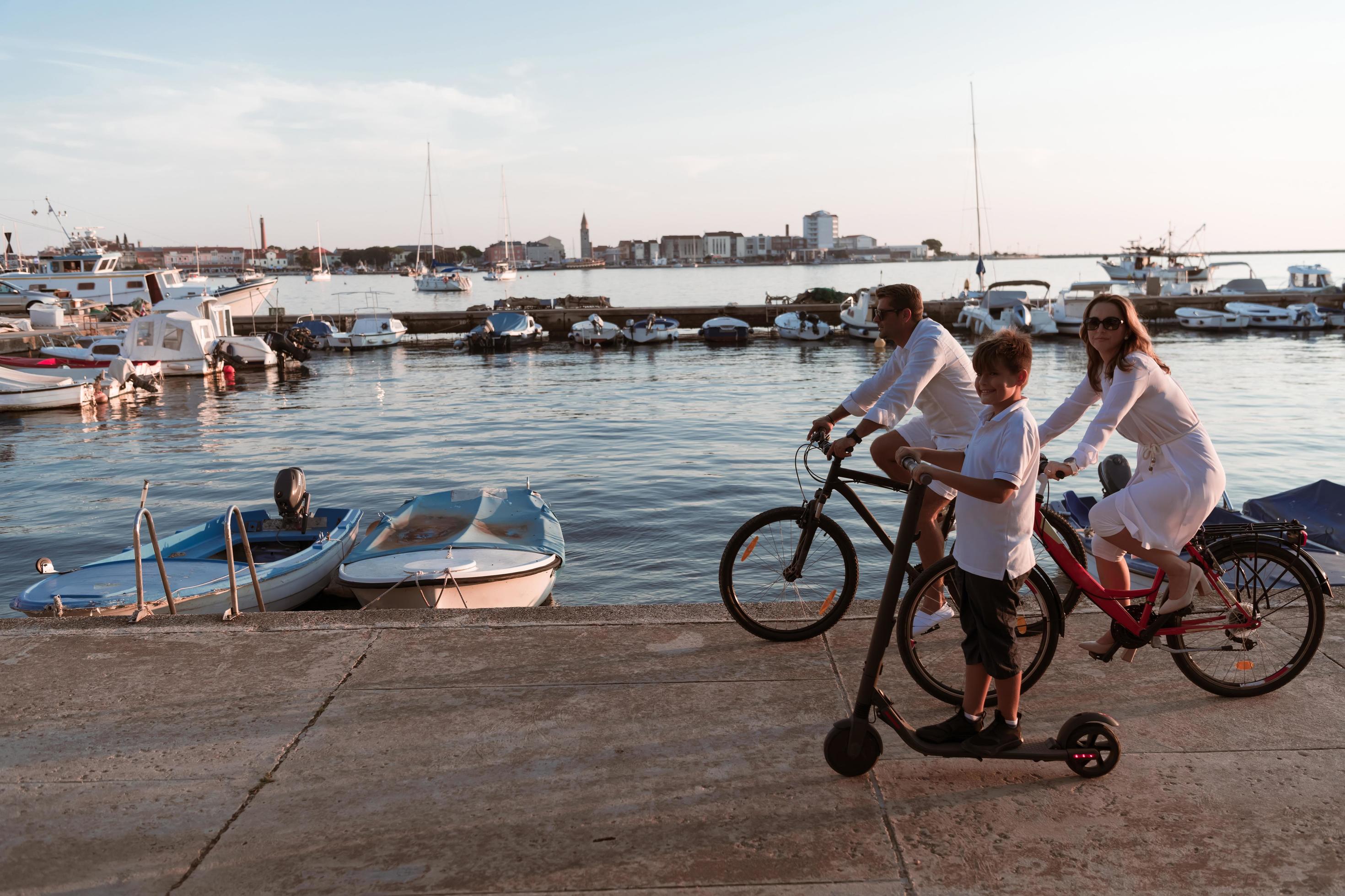 Happy family enjoying a beautiful morning by the sea together, parents riding a bike and their son riding an electric scooter. Selective focus Stock Free