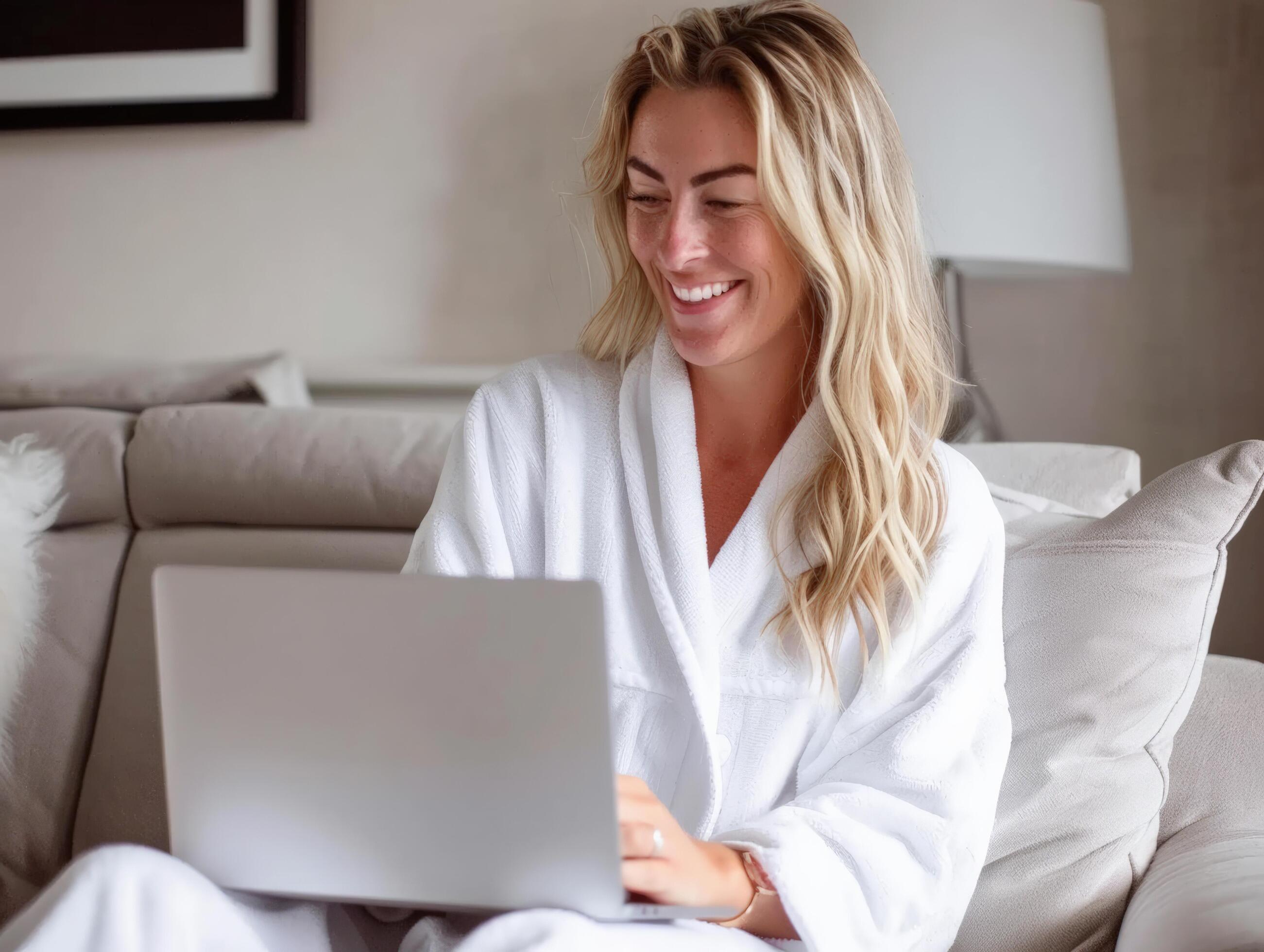 Smiling woman in a white bathrobe using a laptop while seated on a comfortable couch in a cozy living room setting. Stock Free