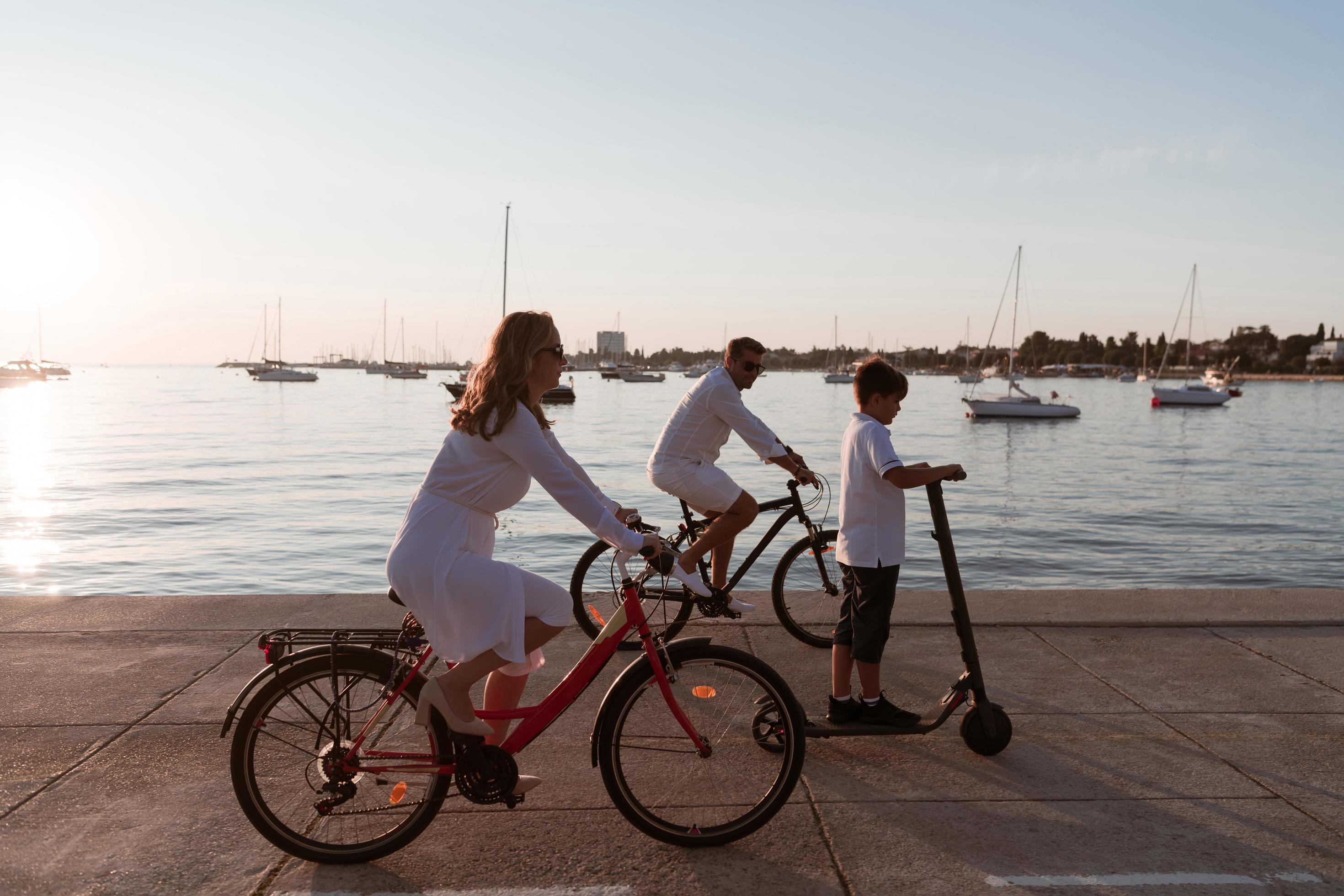 Happy family enjoying a beautiful morning by the sea together, parents riding a bike and their son riding an electric scooter. Selective focus Stock Free