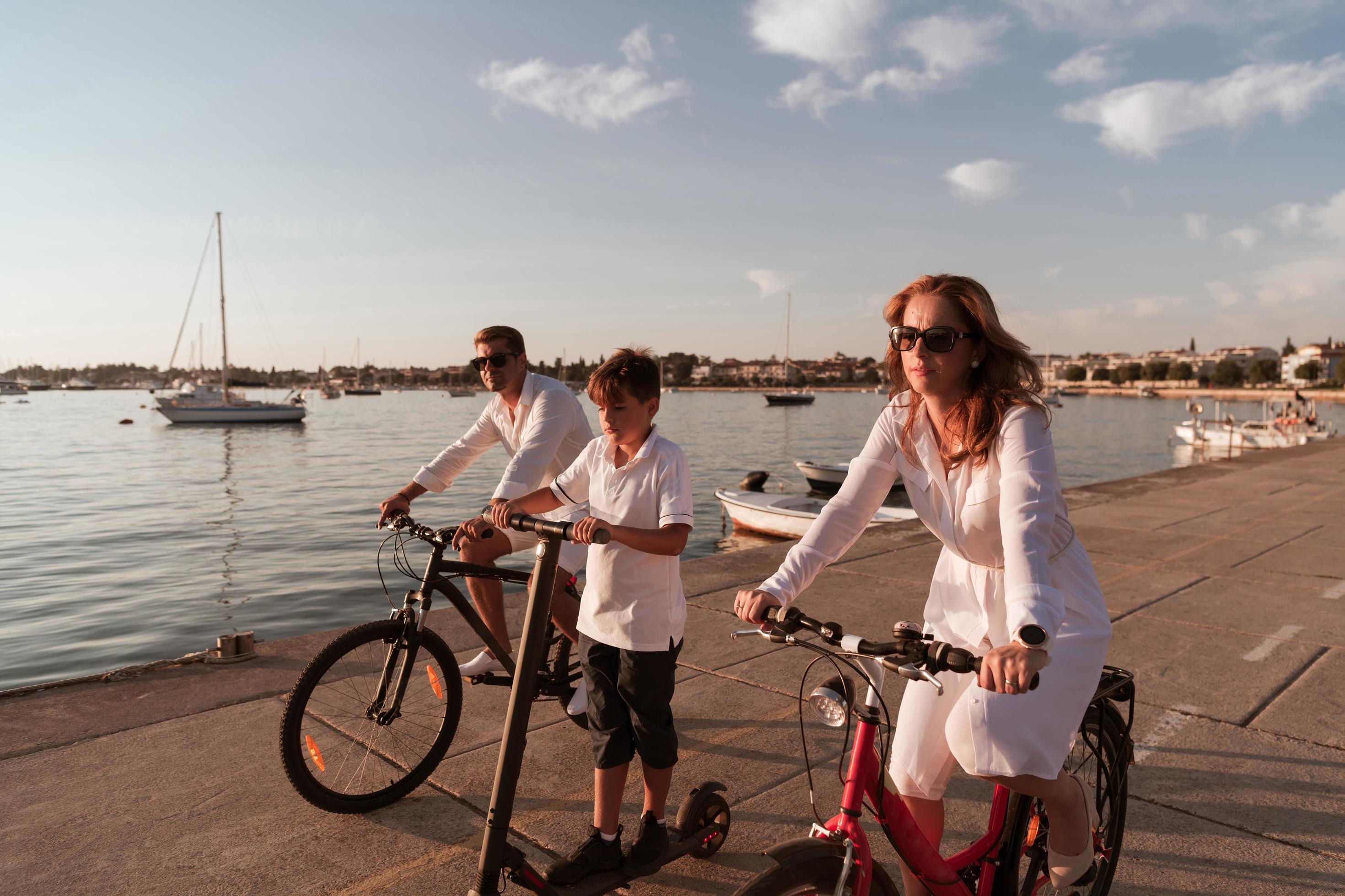 Happy family enjoying a beautiful morning by the sea together, parents riding a bike and their son riding an electric scooter. Selective focus Stock Free