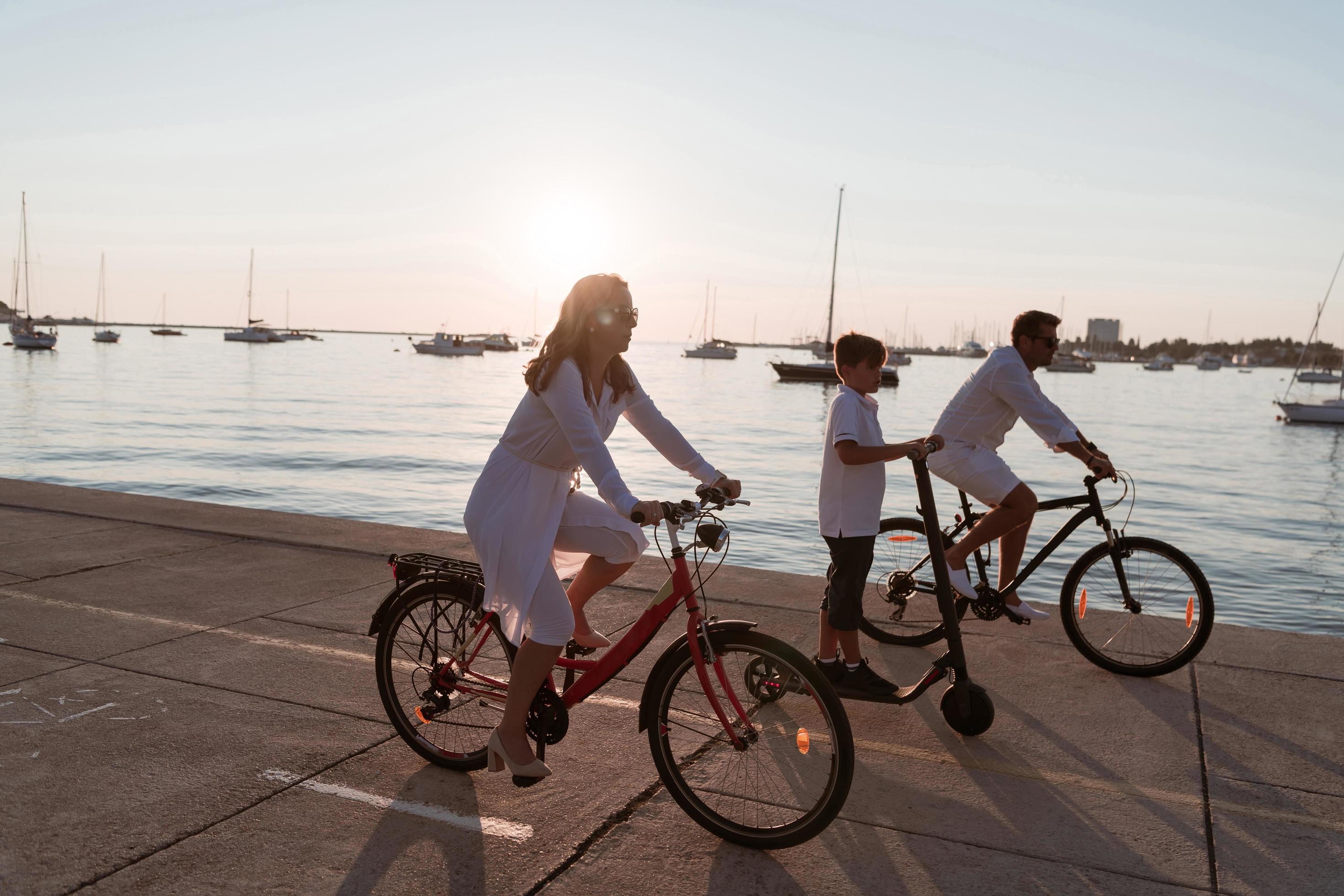 Happy family enjoying a beautiful morning by the sea together, parents riding a bike and their son riding an electric scooter. Selective focus Stock Free