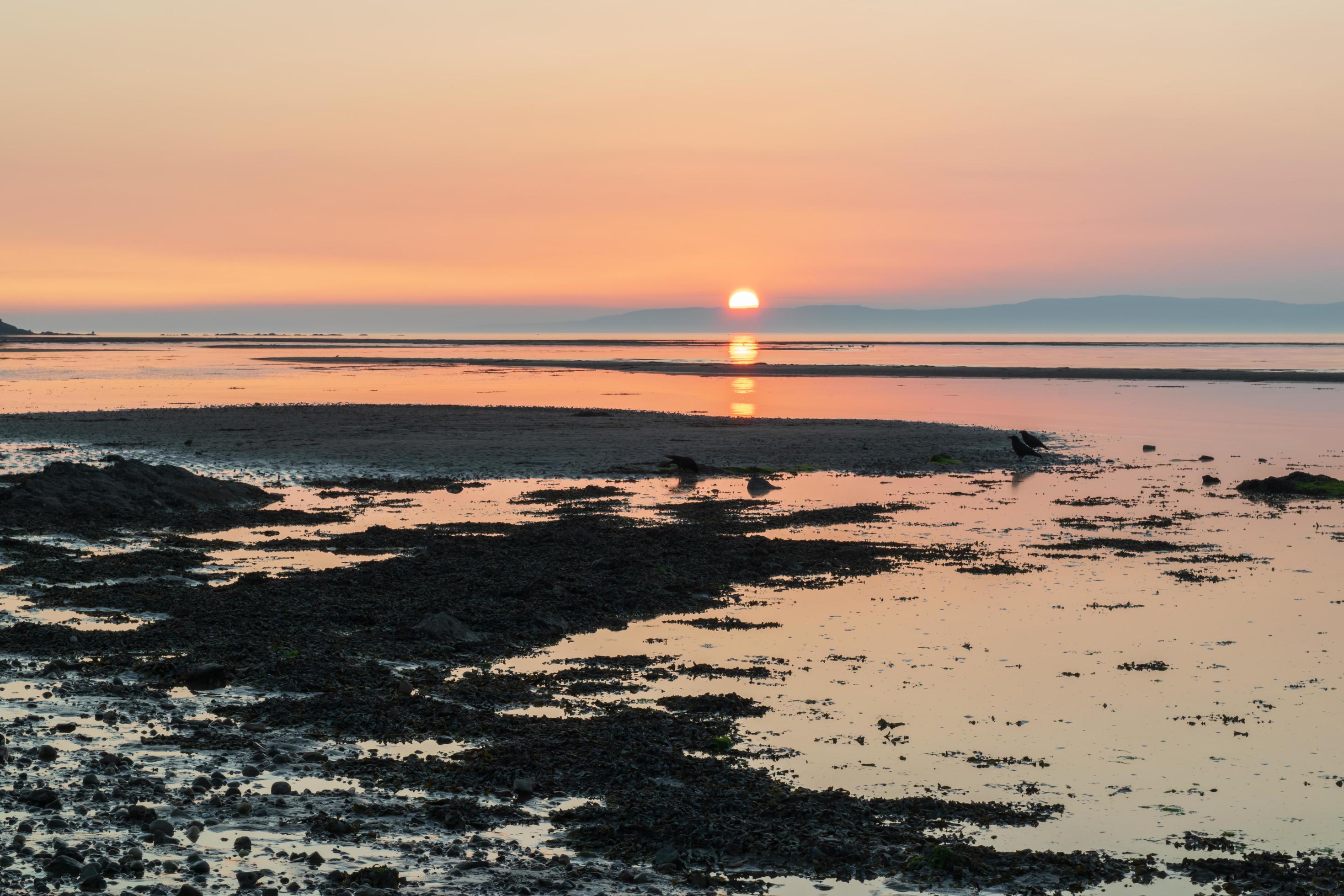 Sunset over the beach behind the mountain. Colourful horizontal line sun reflection on water, mirror. Stock Free