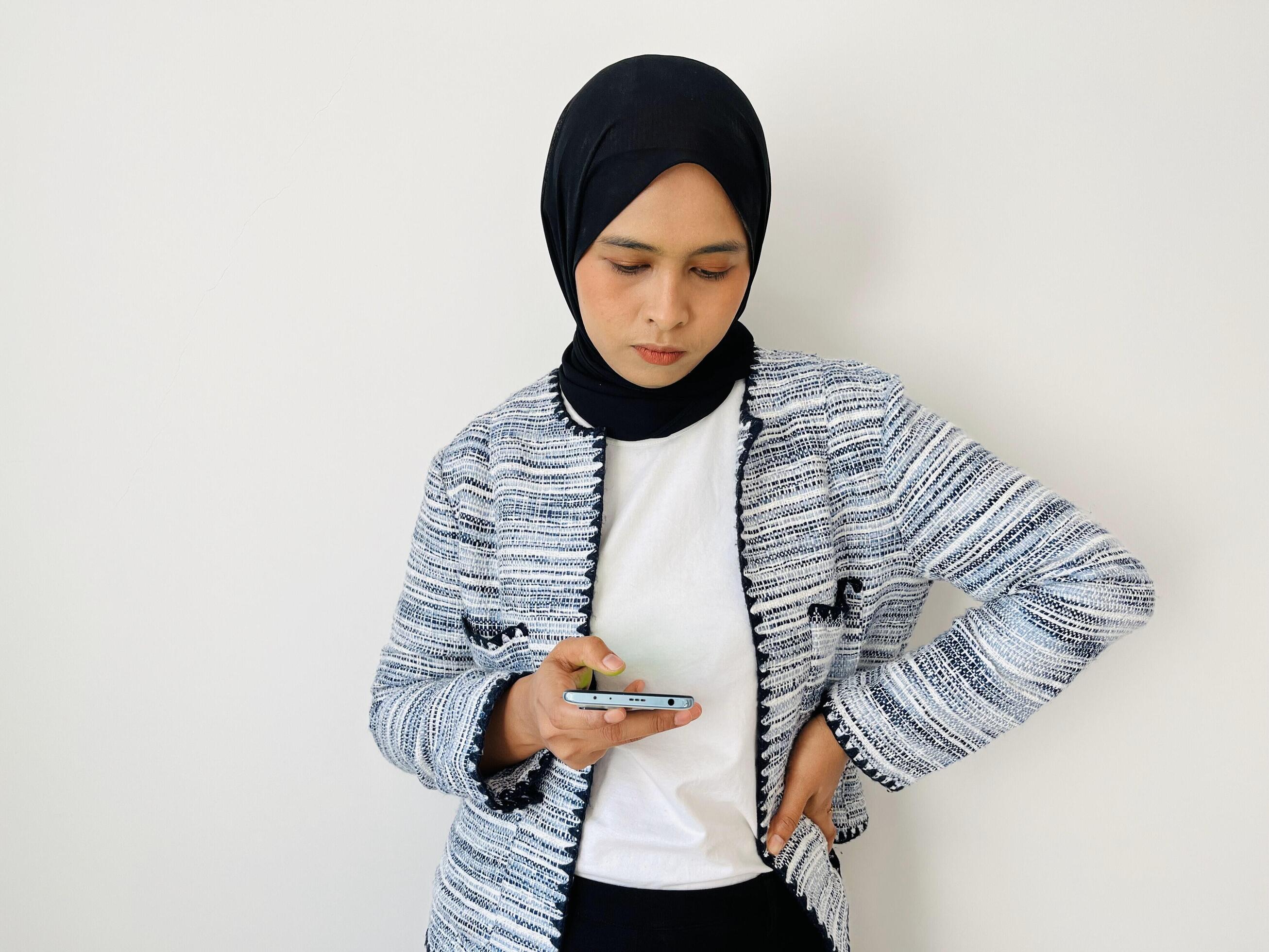 A young Asian Muslim woman holds and looks at a smartphone in annoyance with her other hand on her waist Stock Free