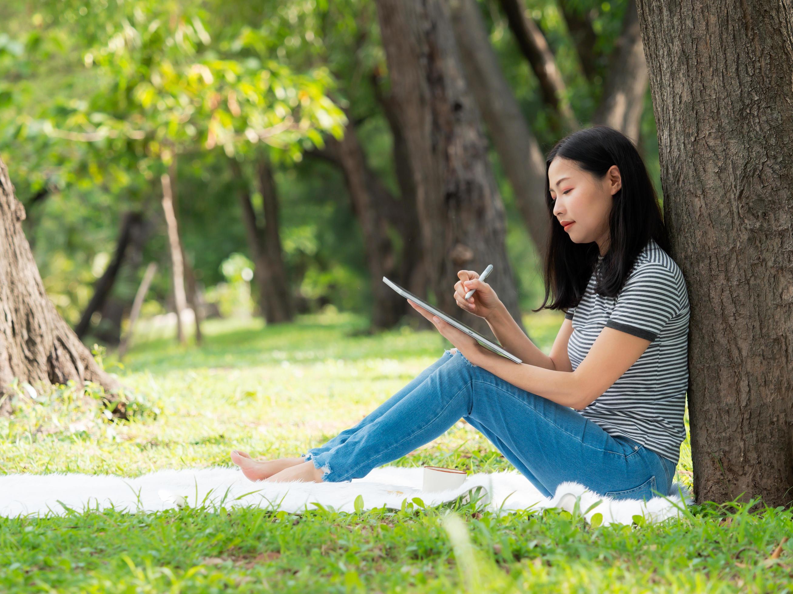 A beautiful Asian woman is sitting in a garden and writing information from the internet onto a digital tablet Stock Free