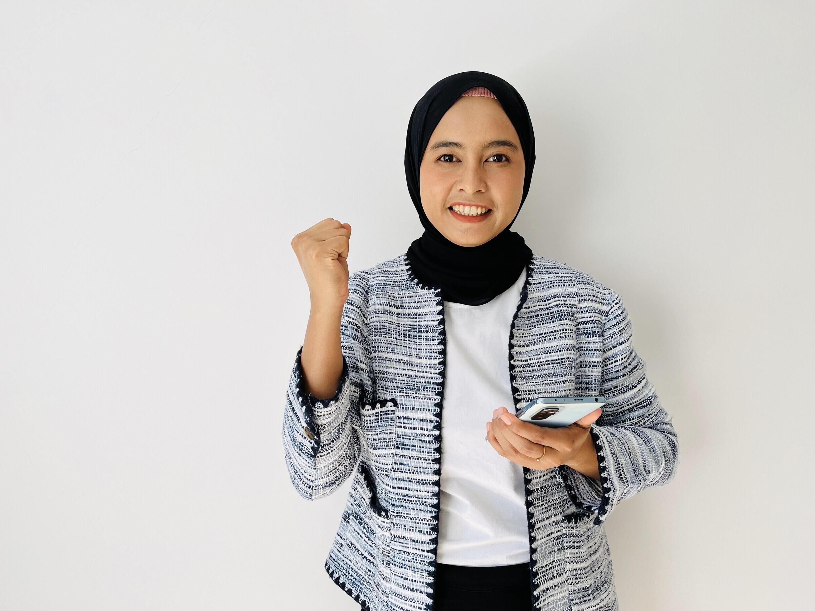 Excited hijab young Asian woman holding mobile phone and gesturing yes, celebrating success, raising clenched fist isolated on white background Stock Free