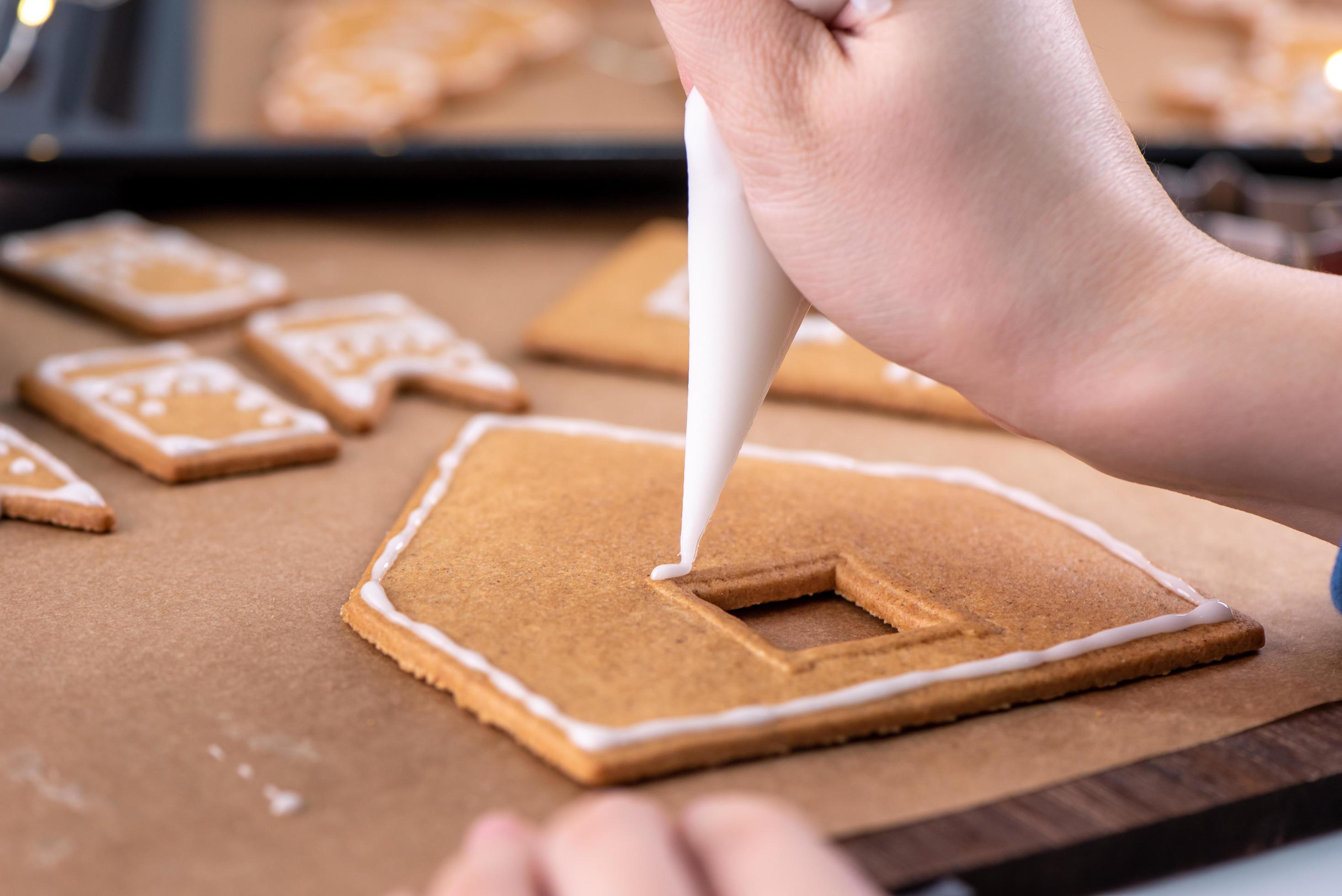 Young woman is decorating Christmas Gingerbread House cookies biscuit at home with frosting topping in icing bag, close up, lifestyle. Stock Free