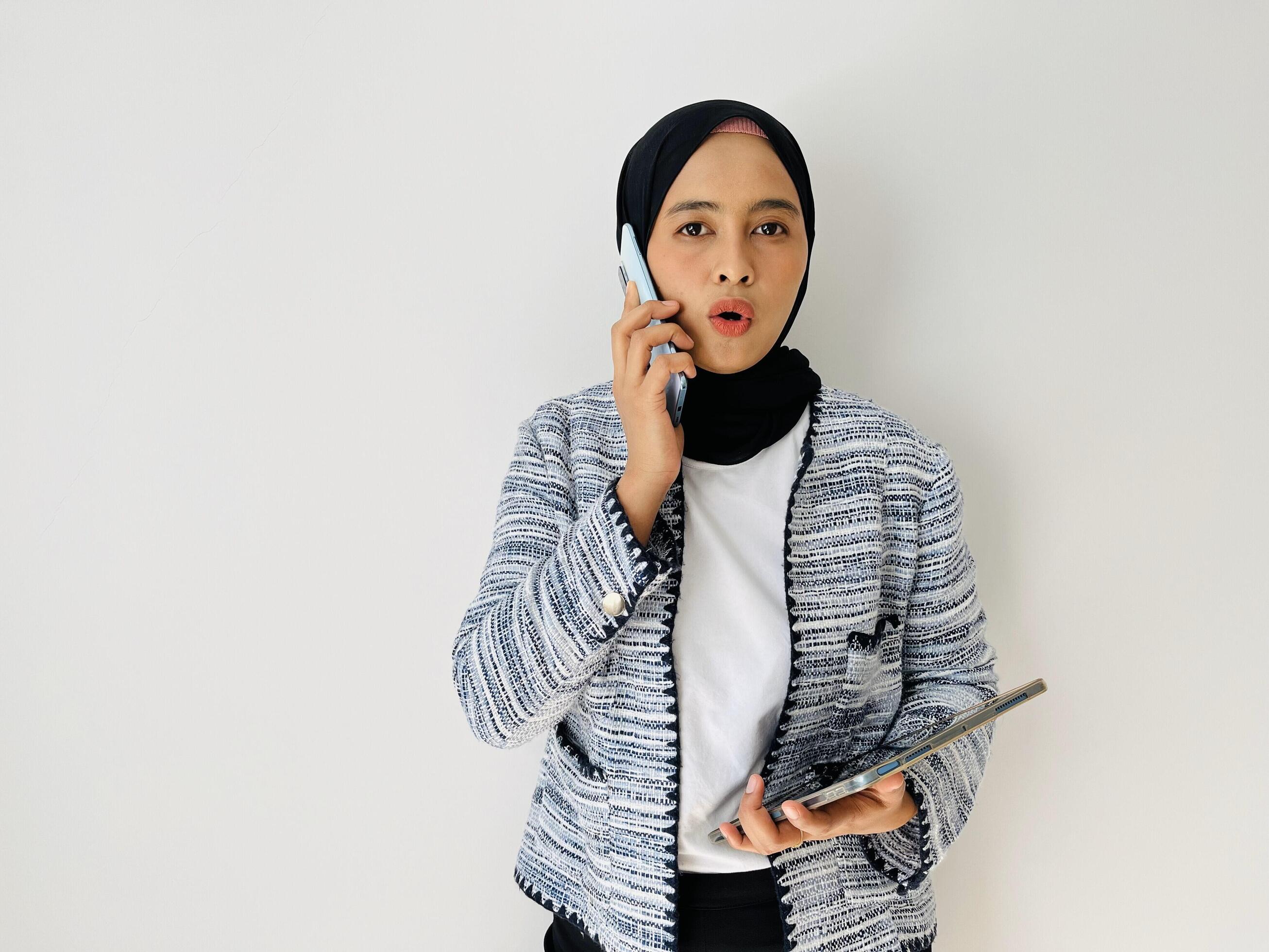 Young Asian woman using mobile phone having call. Standing female wearing black hijab and tweed blazer isolated on white background. Stock Free