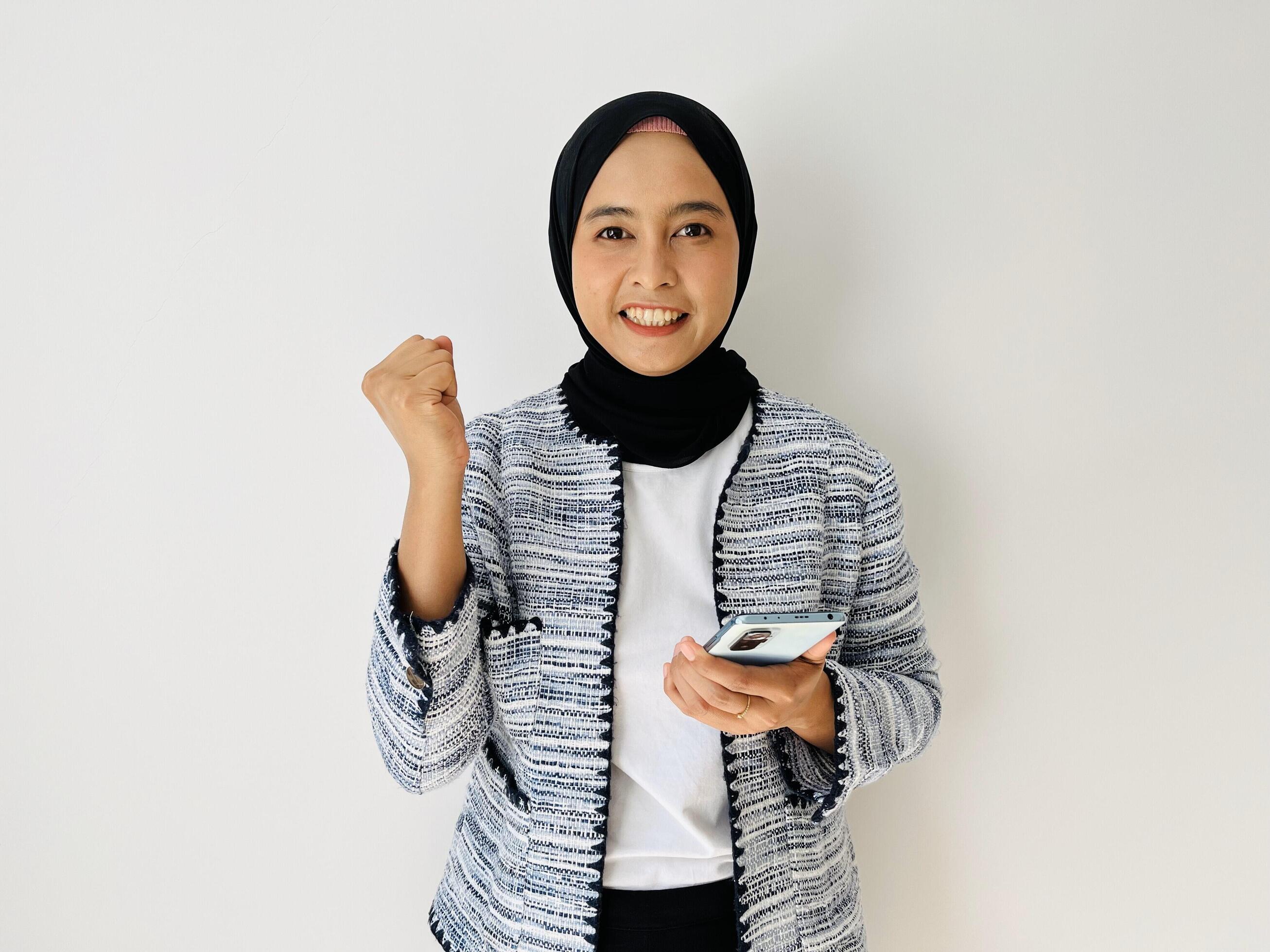 Excited hijab young Asian woman holding mobile phone and gesturing yes, celebrating success, raising clenched fist isolated on white background Stock Free