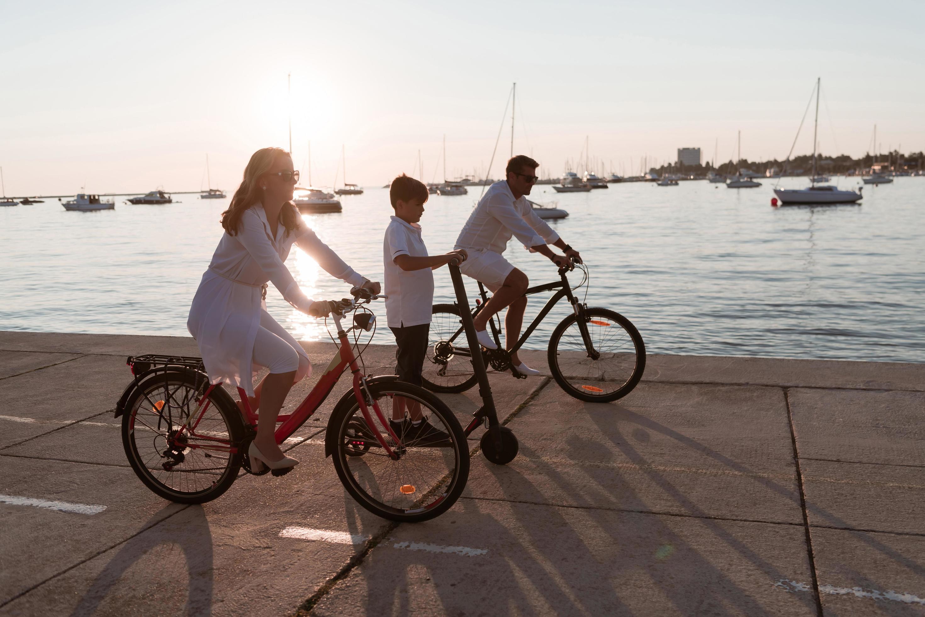 Happy family enjoying a beautiful morning by the sea together, parents riding a bike and their son riding an electric scooter. Selective focus Stock Free